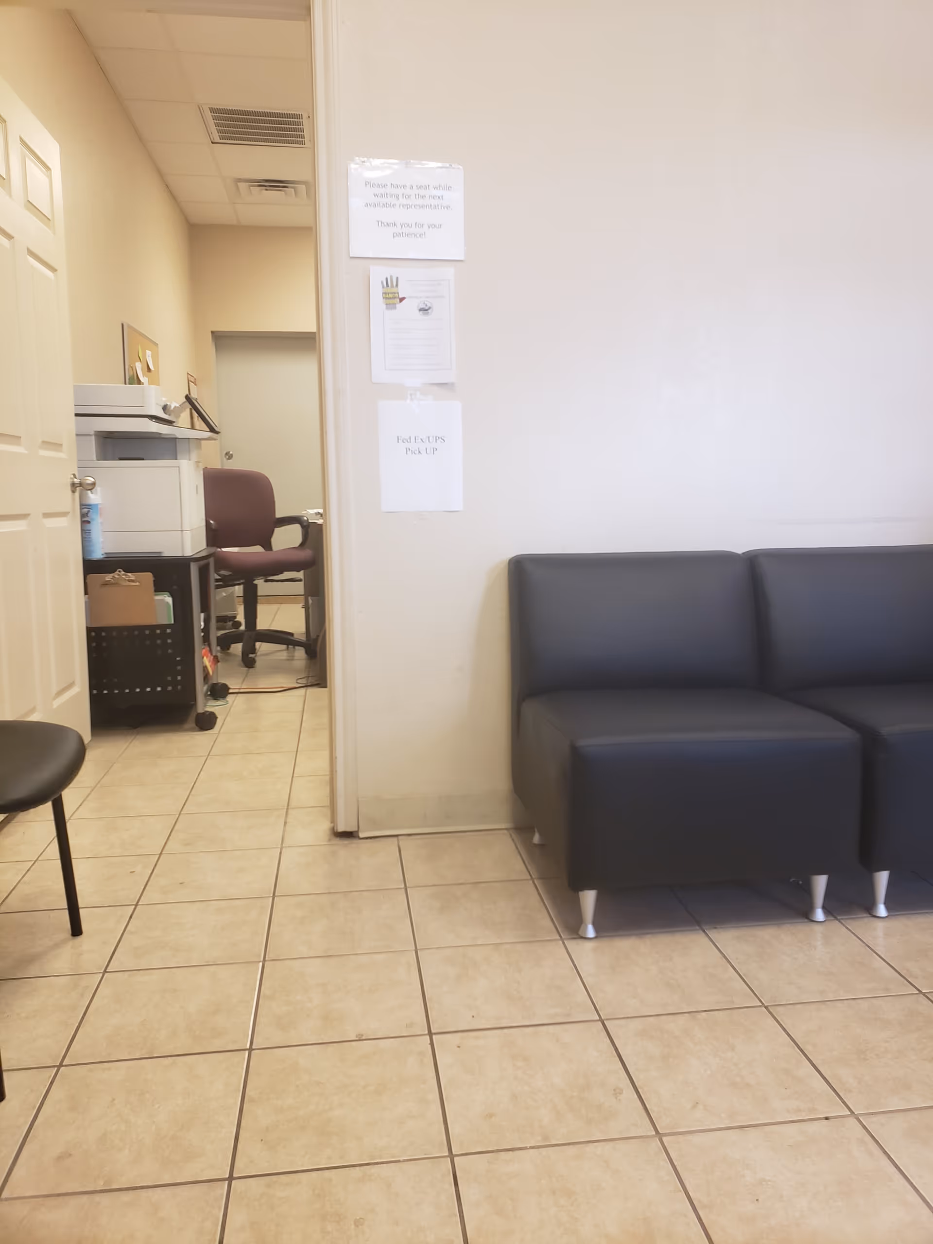 Empty waiting area with a black sofa, tiled floor, and hallway leading to an office.