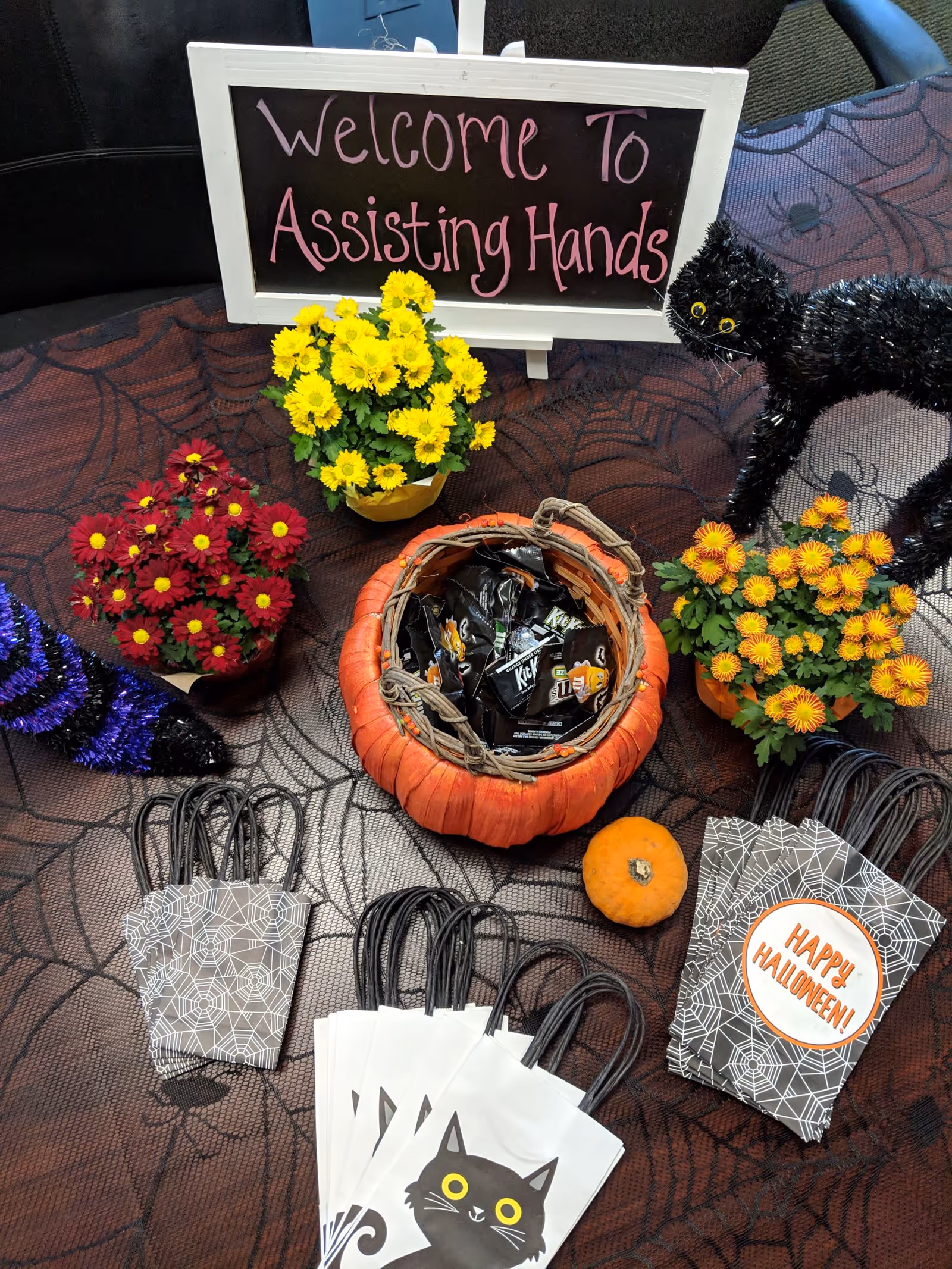 Halloween decor on a table with flowers, a pumpkin basket, and a sign reading Assisting Hands.