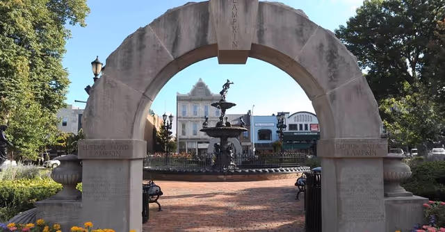 Outdoor plaza featuring a large stone arch, fountain, brick walkway, and surrounding storefronts.