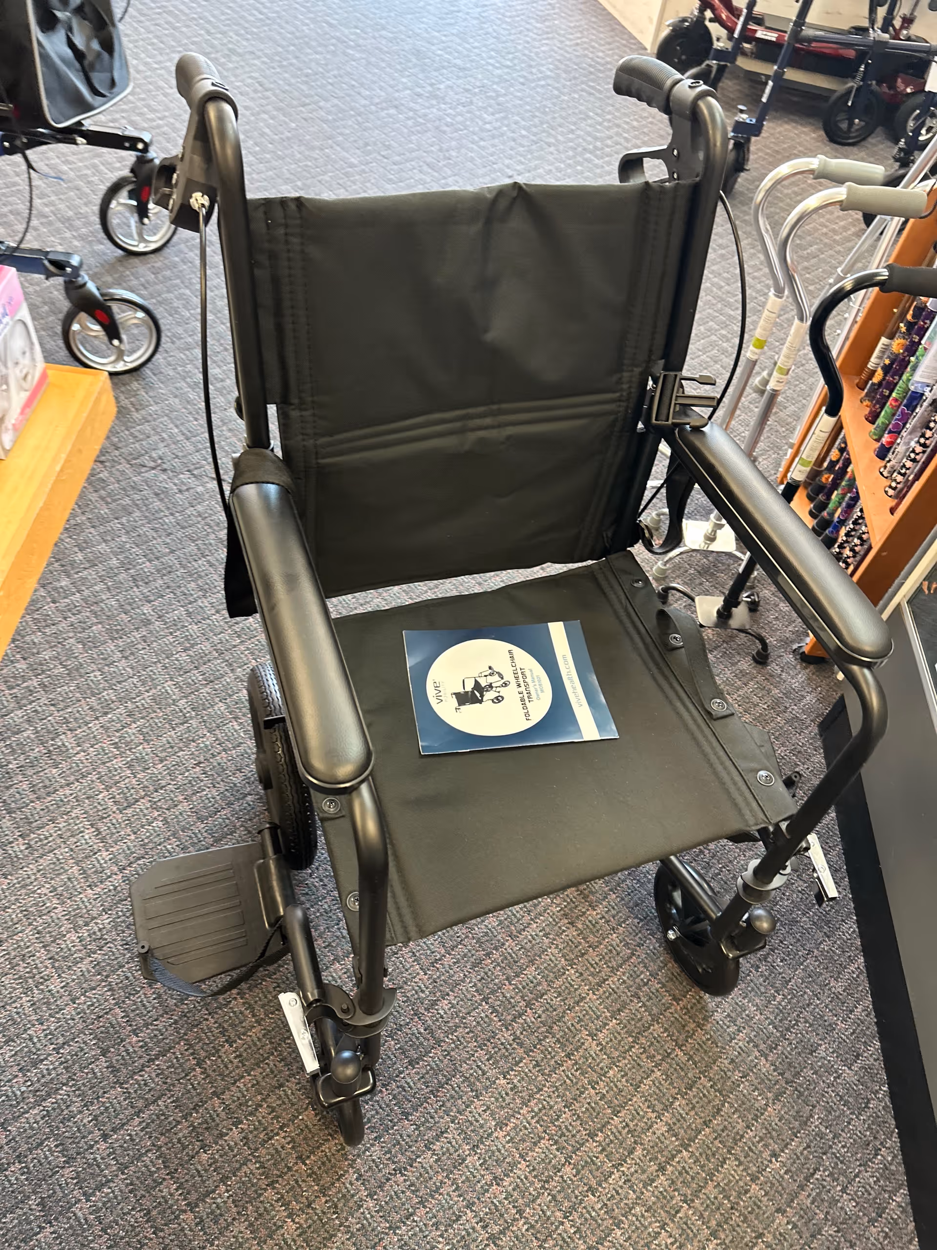 Black wheelchair sits on a carpeted showroom floor among medical supplies.