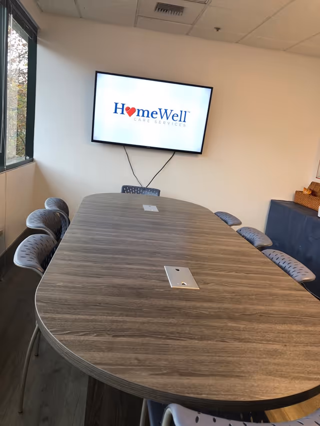 Oval conference table with gray chairs in a room featuring a wall-mounted HomeWell Care Services screen.