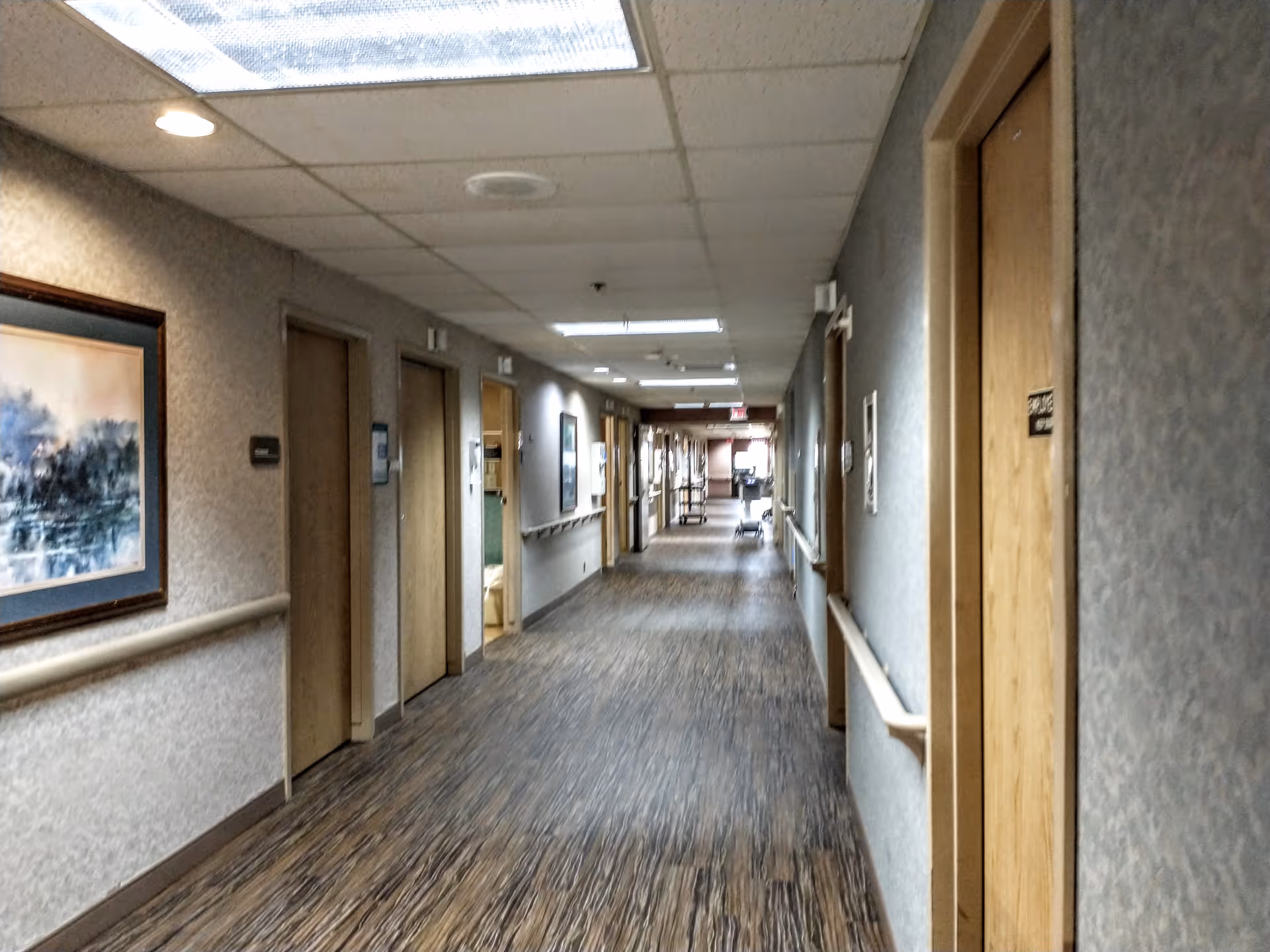 Long hospital-like hallway with doors, wall rails, and overhead lights in a senior living facility.