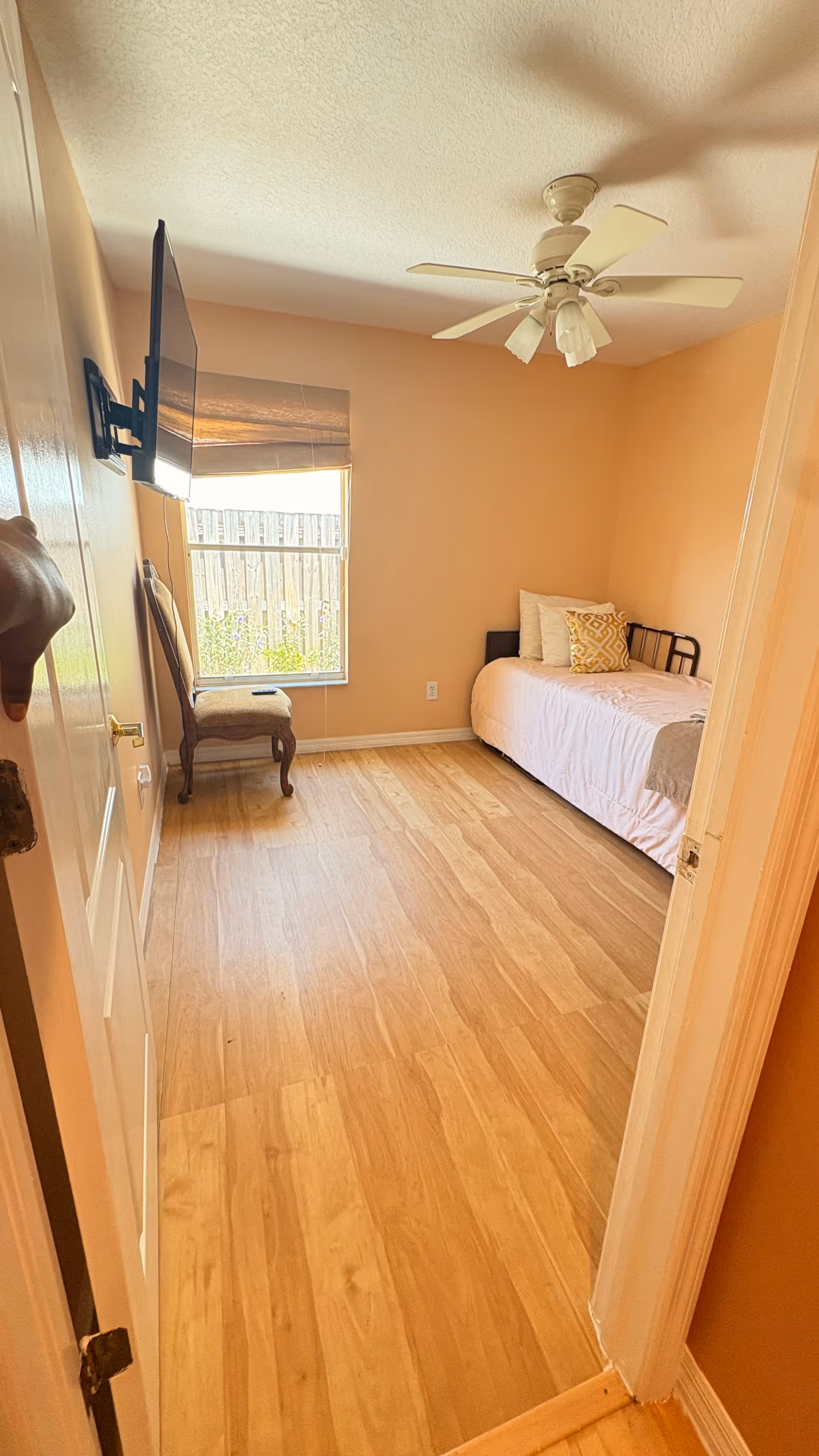 Peach-colored bedroom with a bed, ceiling fan, wall-mounted TV, window, and chair.
