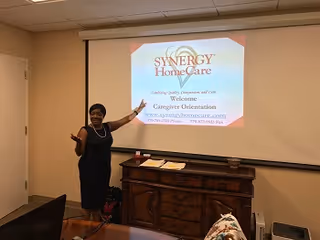 Presenter standing by projector screen displaying SYNERGY HomeCare logo in a meeting room.