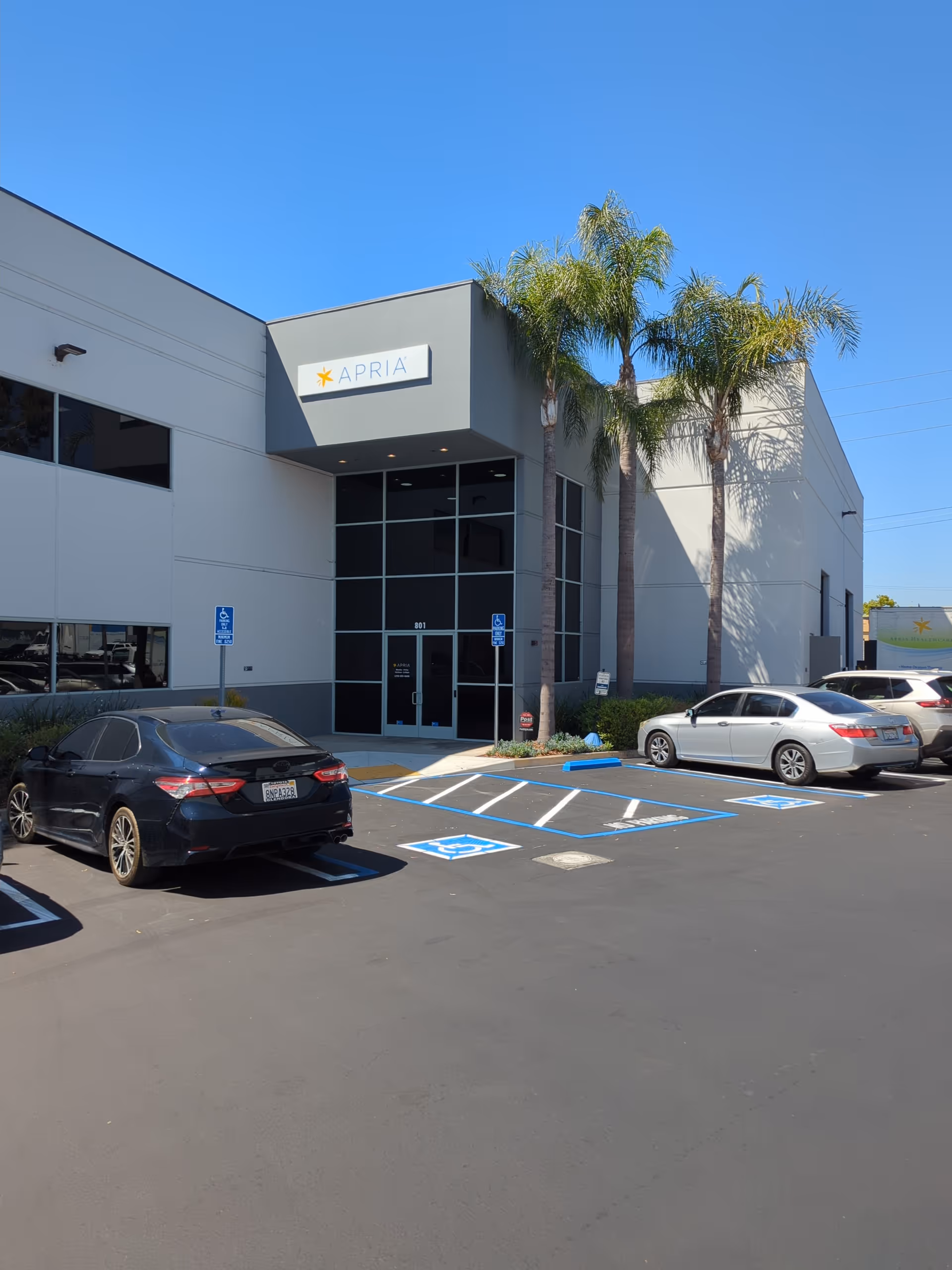 Apria Healthcare exterior with glass entrance, palm trees, and accessible parking in a sunny lot.