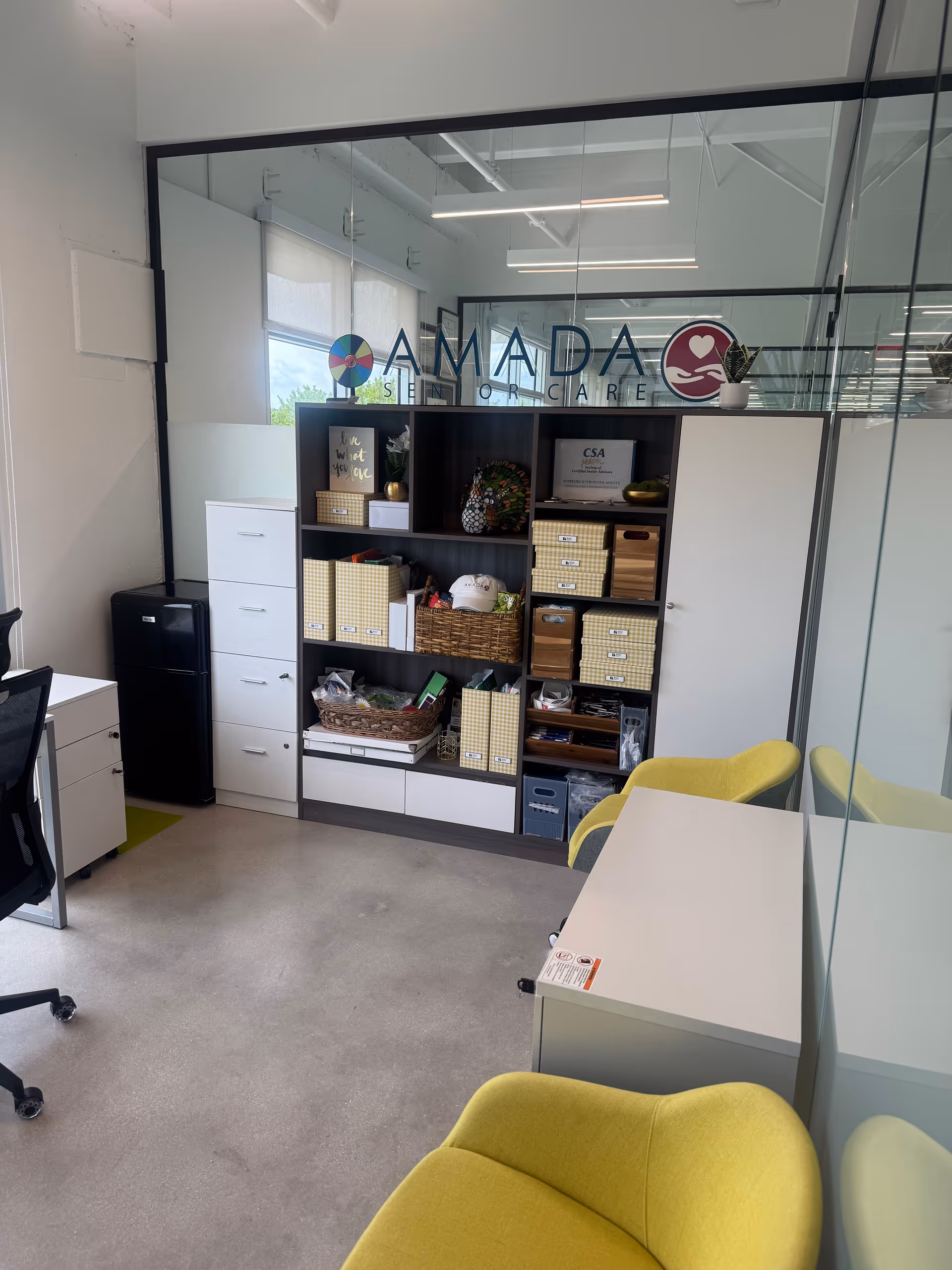 Interior office area of Amada Senior Care with glass partition, shelves, and yellow chairs.