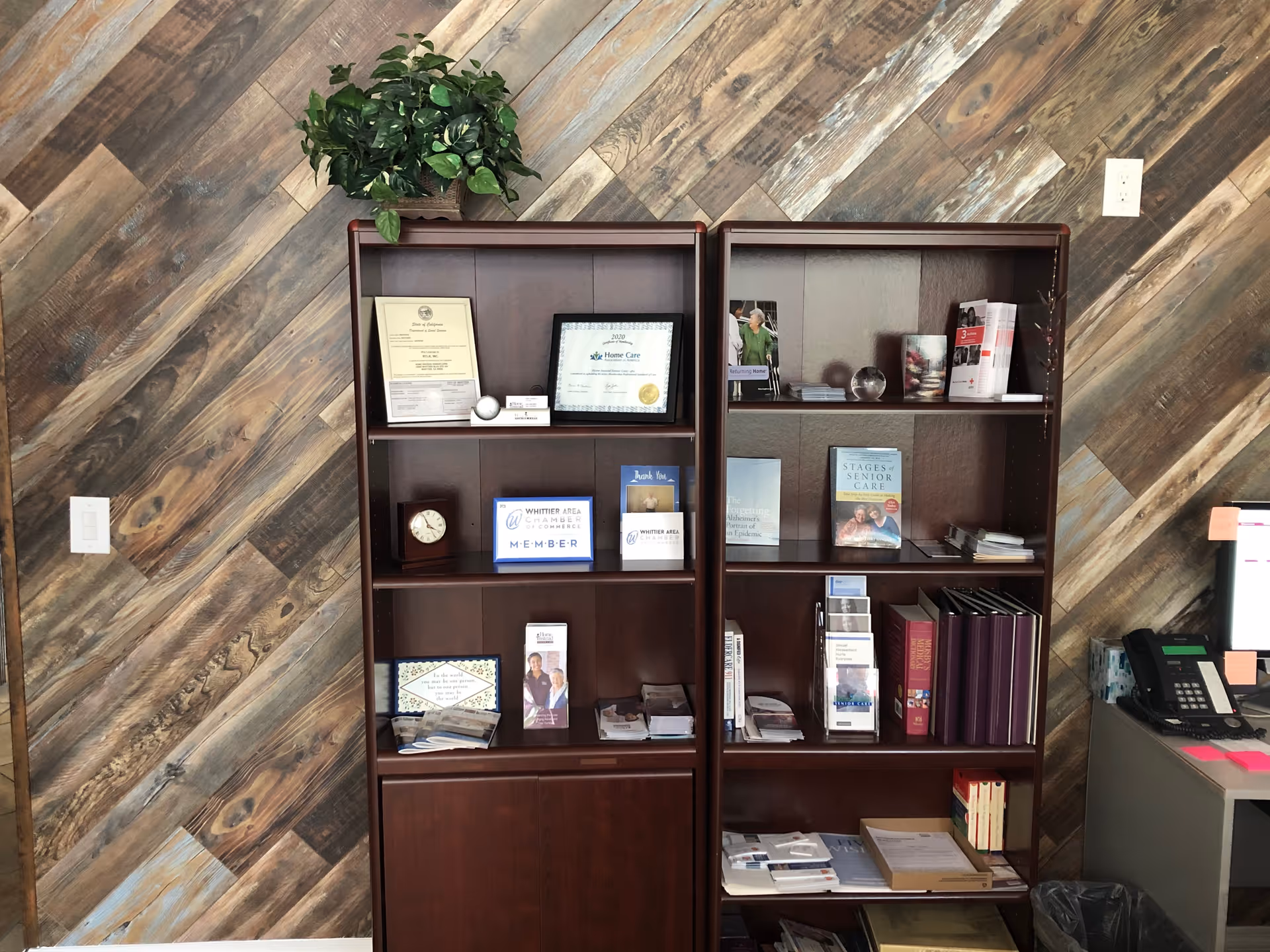 Two dark wood bookshelves filled with brochures and certificates against a diagonal wood wall.