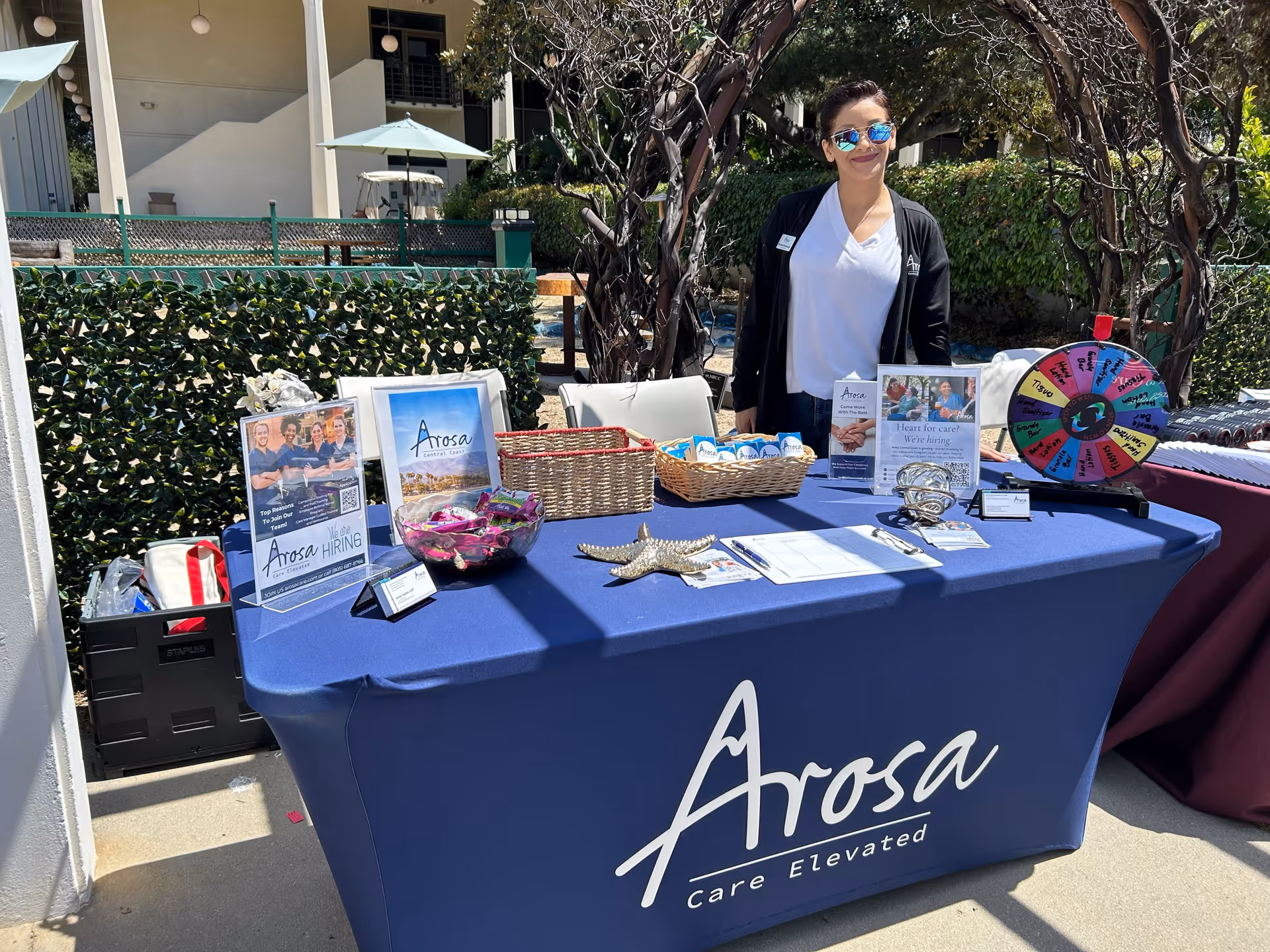 Outdoor promotional booth with a staff member behind a blue Arosa table in a sunny courtyard.