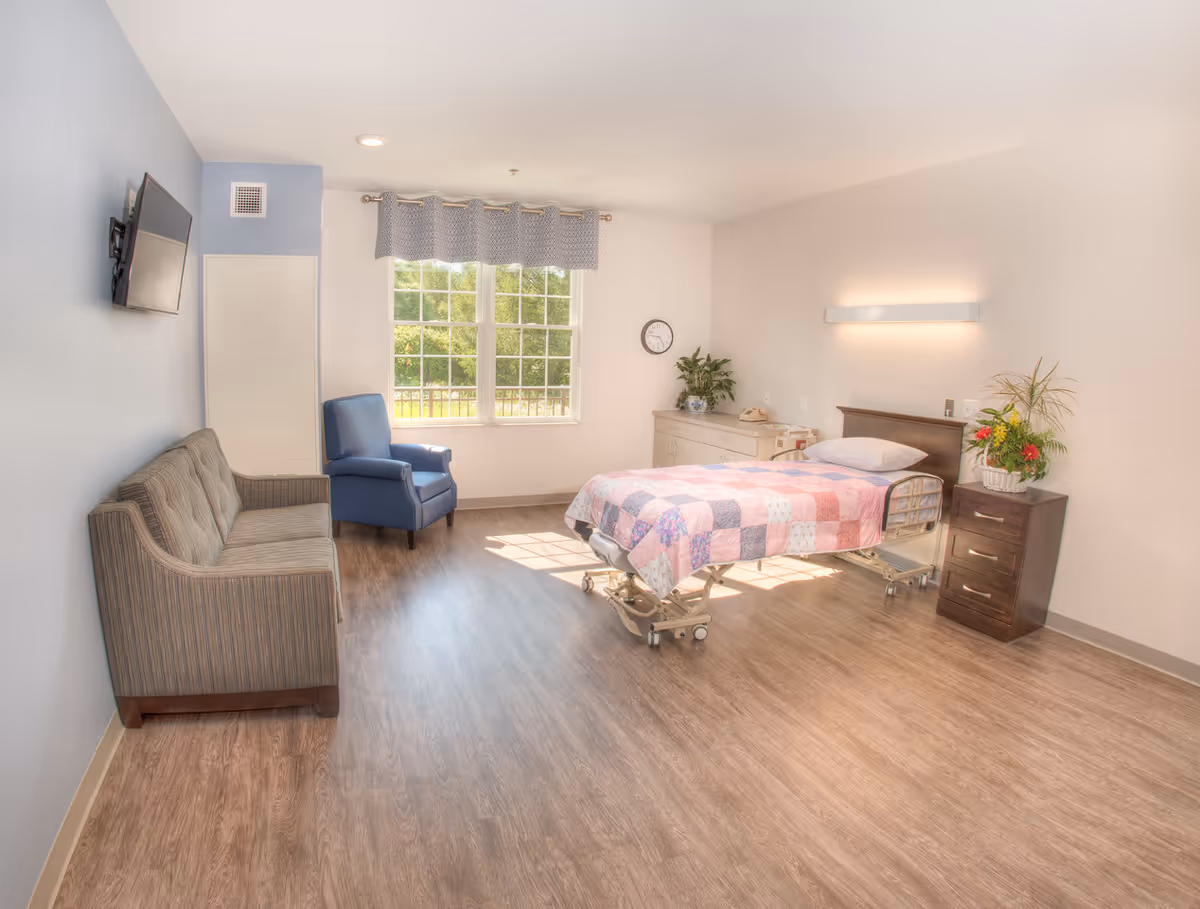 Bright patient room with adjustable bed, quilt, dresser, sofa, blue chair, window, and plants.