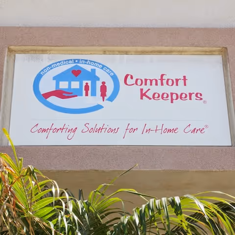 Exterior wall with a Comfort Keepers sign and lush tropical plants in front.