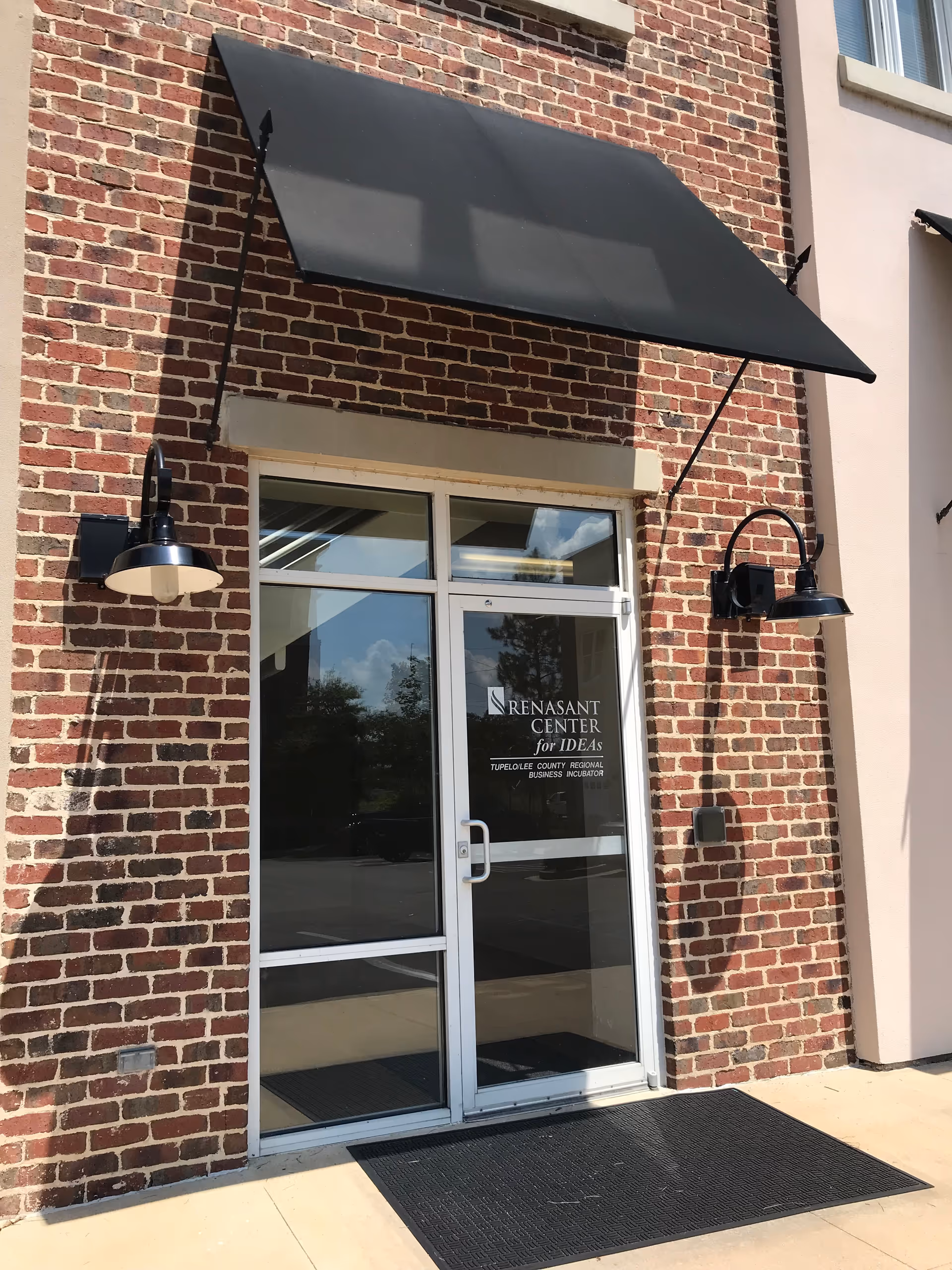 Brick building entrance with glass door, black awning, and exterior lamps.