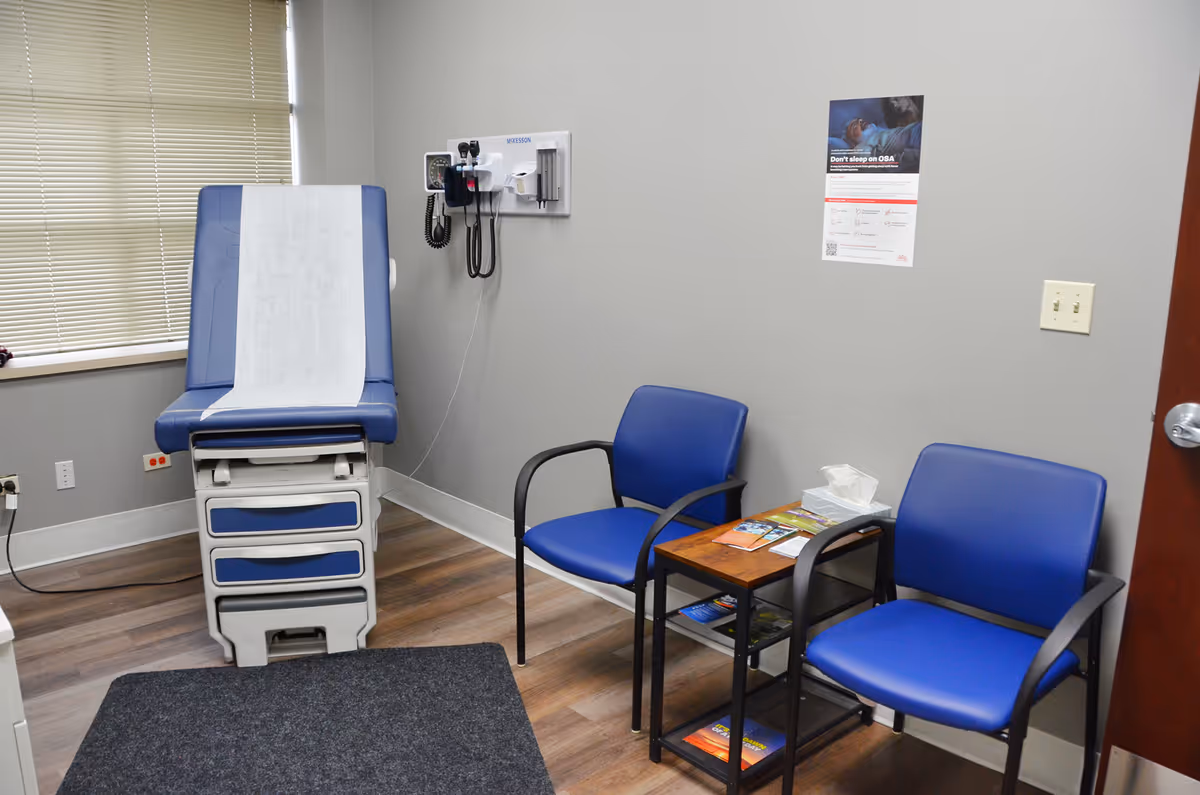 Examination room with blue exam chair, waiting chairs, and medical devices on the wall.