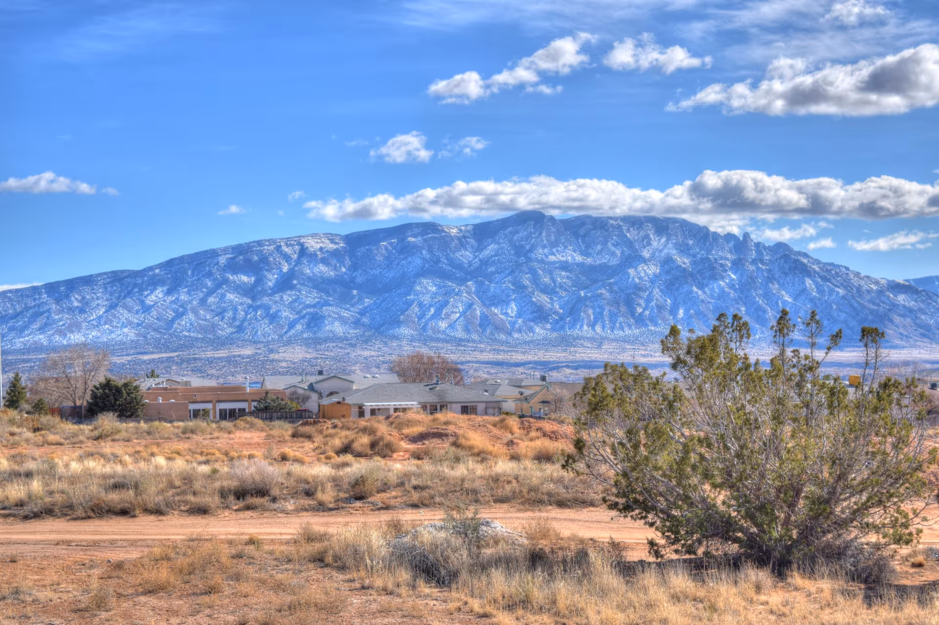 Desert landscape with distant houses and snow-capped mountains under blue sky.