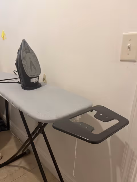 A compact ironing board with a plugged-in iron stands by a wall.