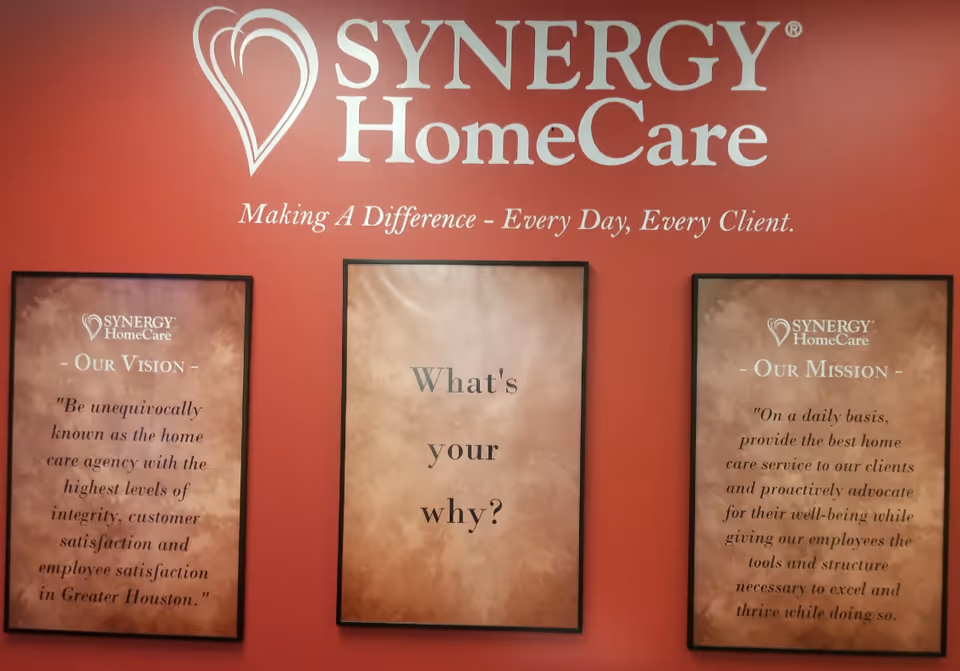 Red wall displays SYNERGY HomeCare logo and three framed posters.