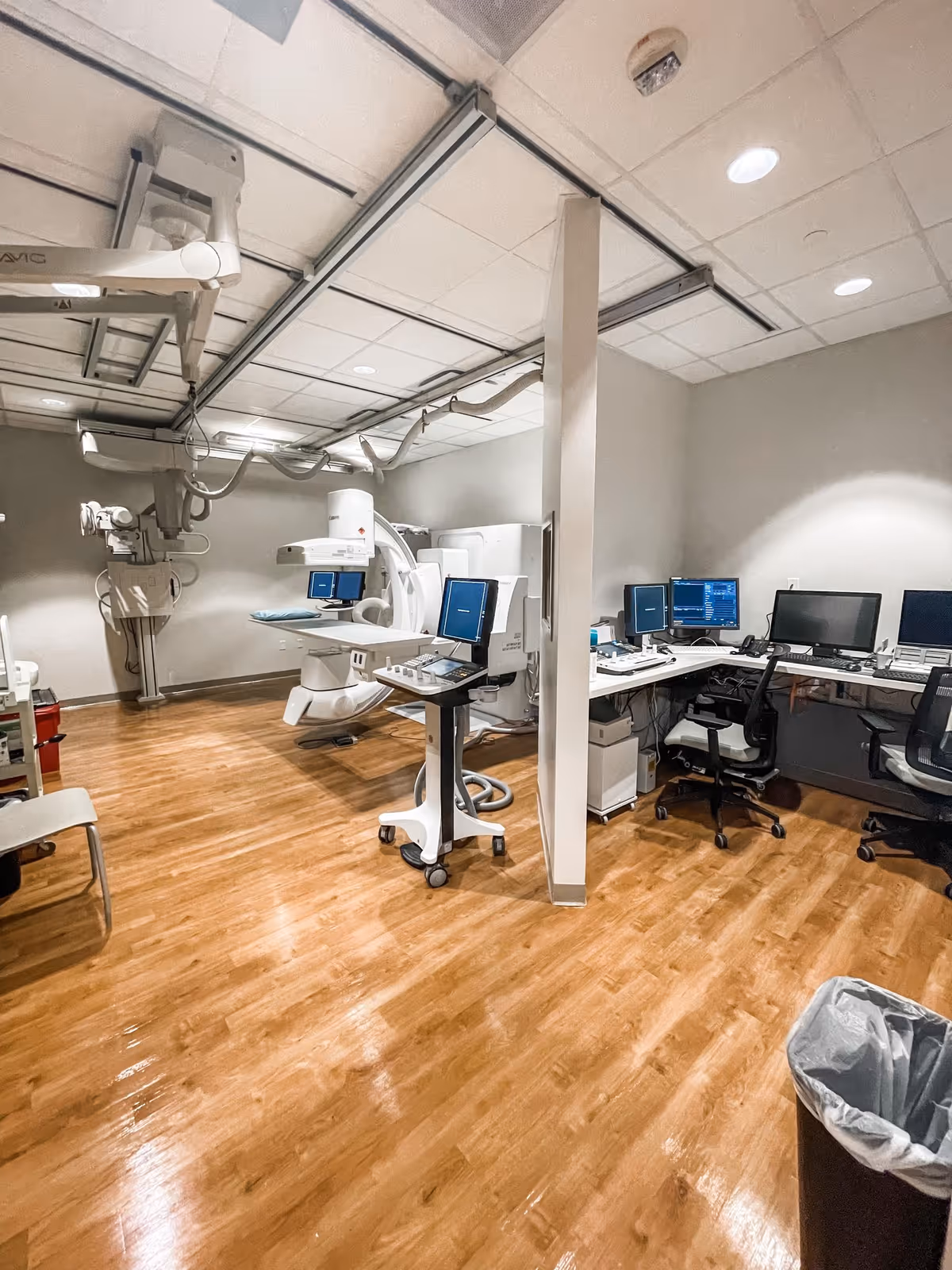 Medical imaging room with C-arm equipment, monitors, and desks in a clinical interior.