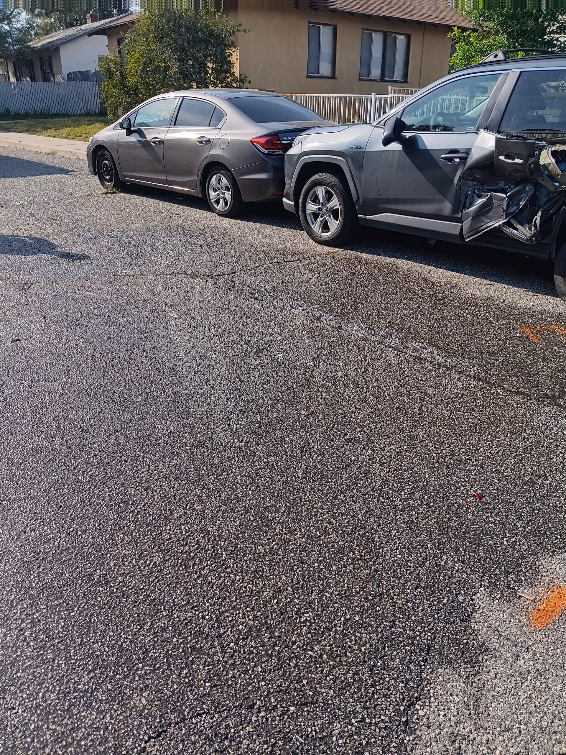 Two damaged cars parked on a residential street in front of a beige house.