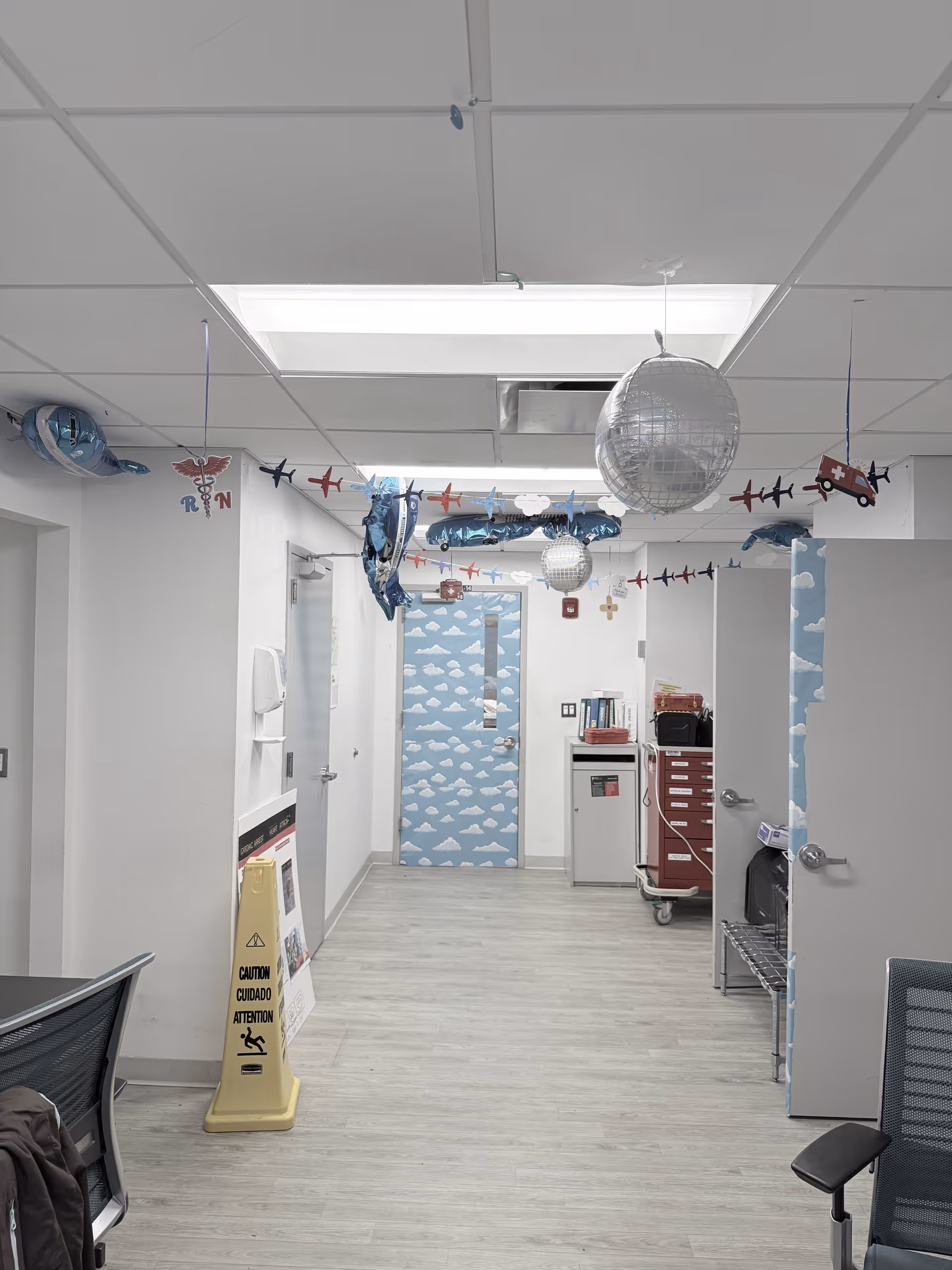 Interior hospital-like hallway with ceiling tiles, garlands, cloud wallpaper doors, and a medical cart.