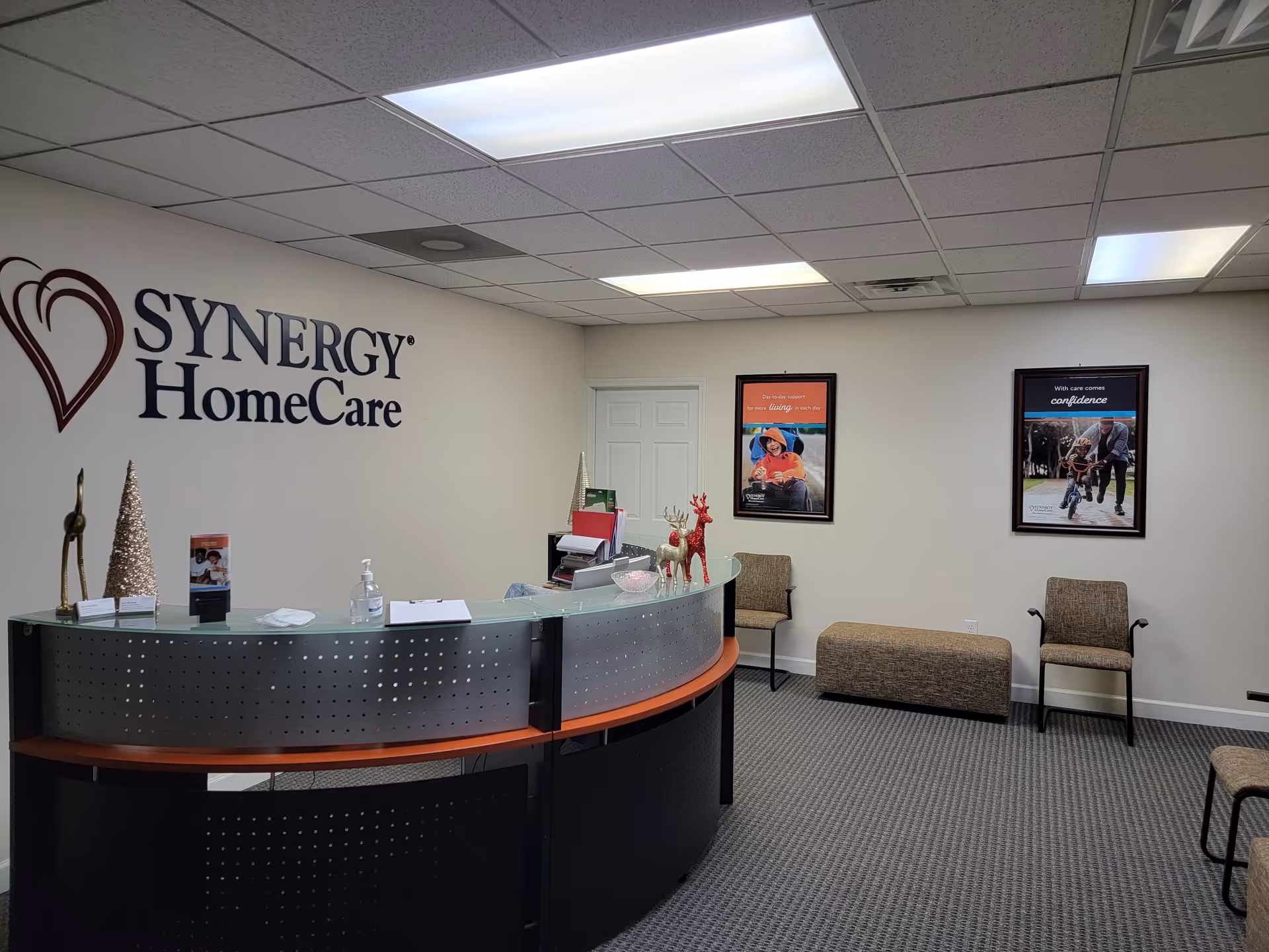 Reception area with SYNERGY HomeCare logo, curved desk, framed posters, and seating.