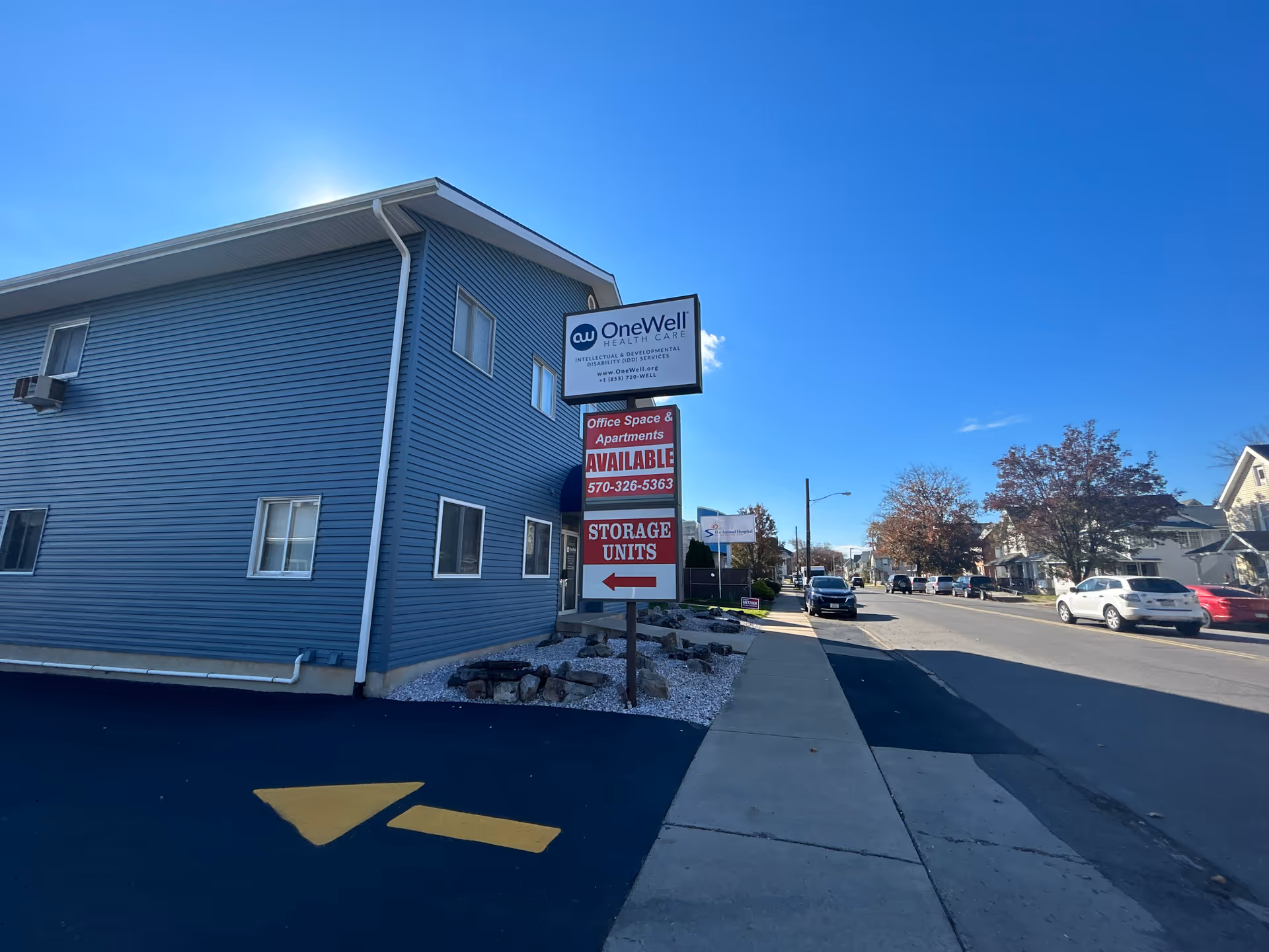 Blue two-story OneWell Health Care building with sidewalk, street, and parked cars.