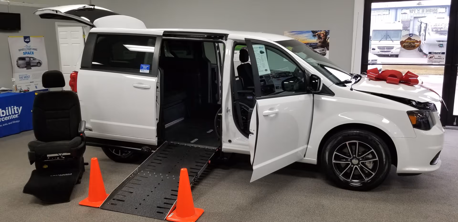 White wheelchair-accessible van with a deployed ramp, open doors, and a red bow inside showroom.