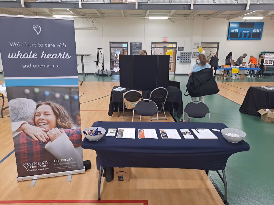 Indoors at a community event, a SYNERGY HomeCare booth with banner and brochures.