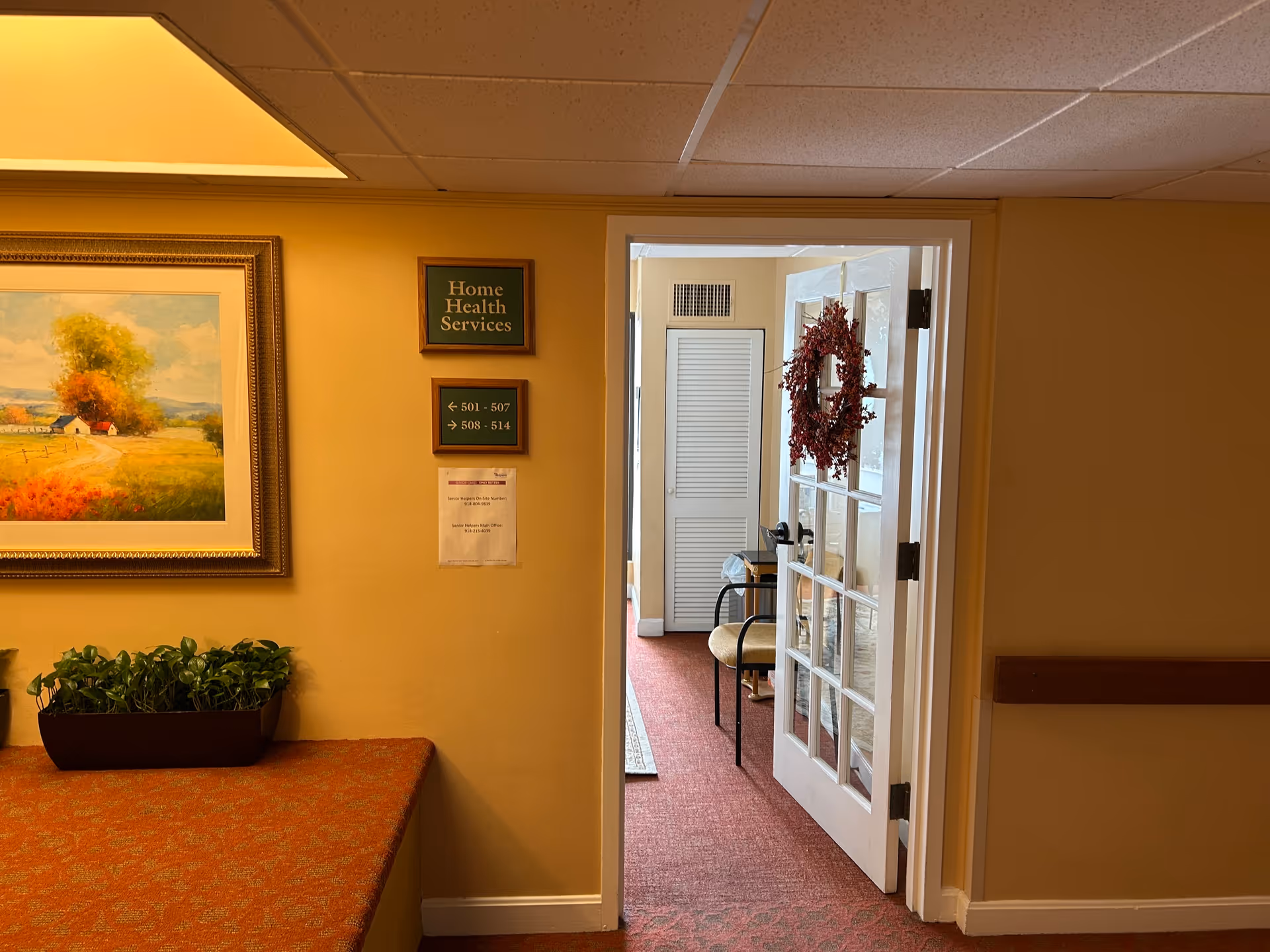 Interior hallway of a senior living facility with a door, wreath, framed artwork, and potted plants.