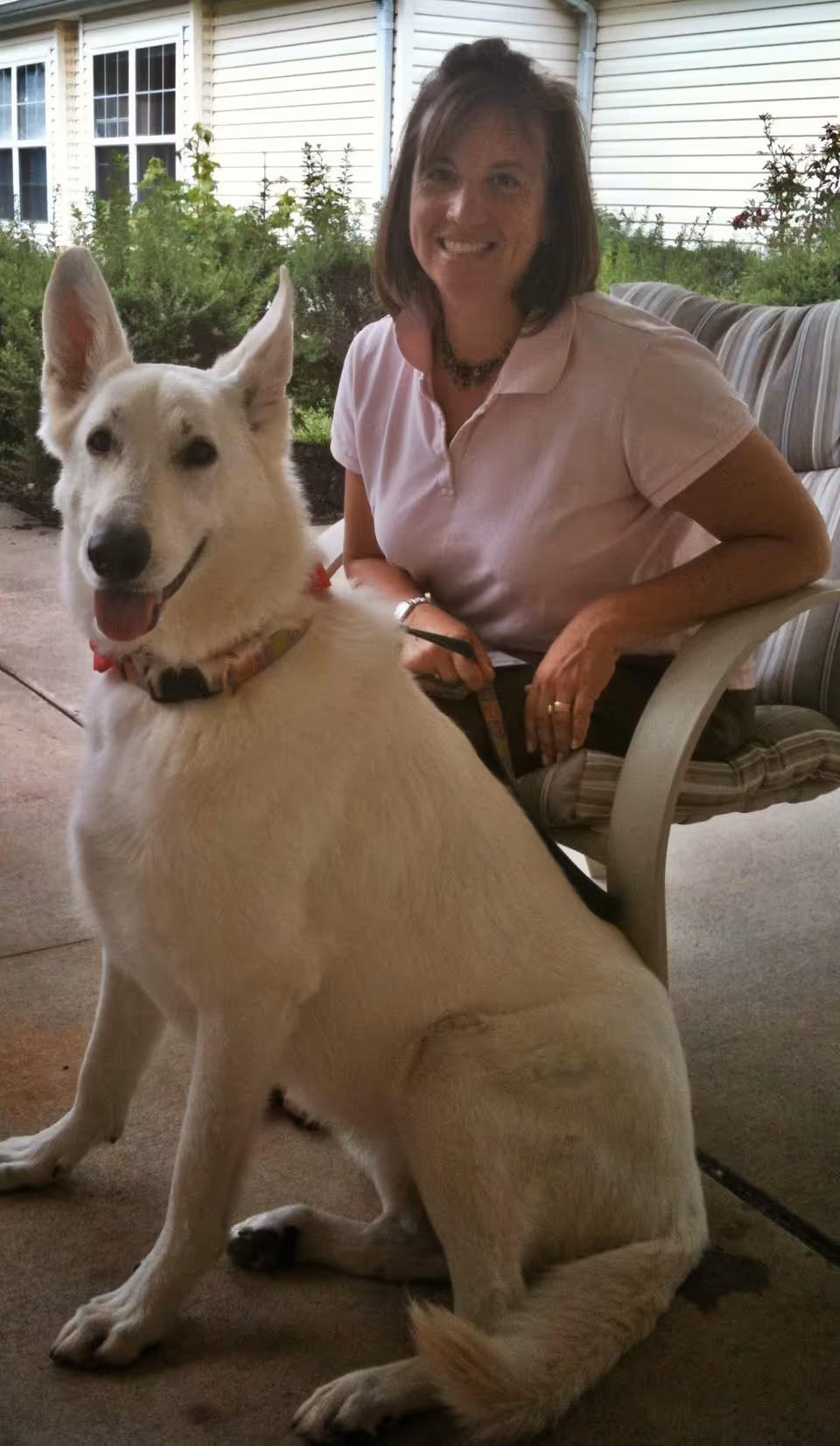 A smiling woman sits on an outdoor patio with a white dog beside her.