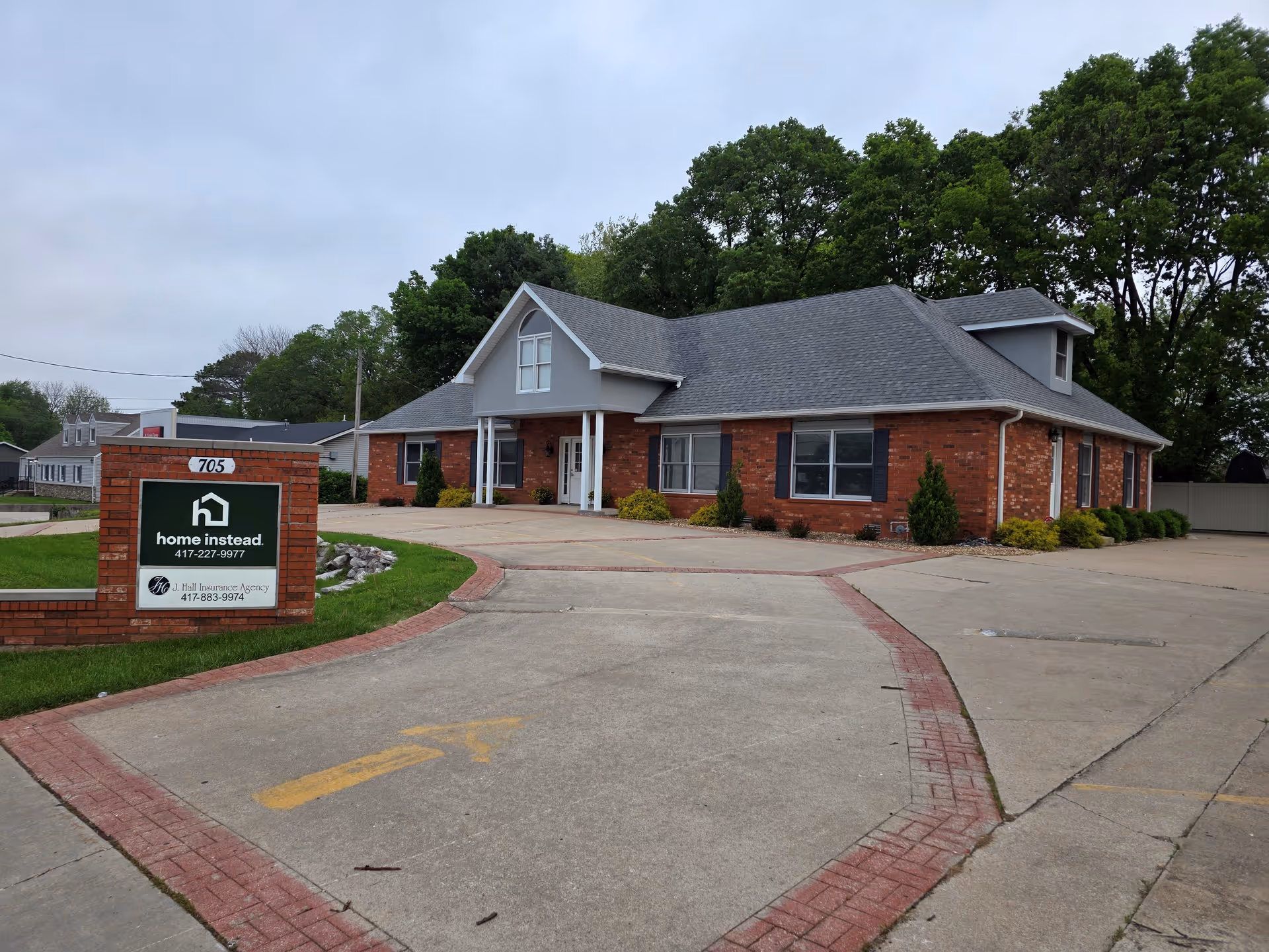 Brick senior living facility with curved driveway, sign reading Home Instead, and manicured shrubs.