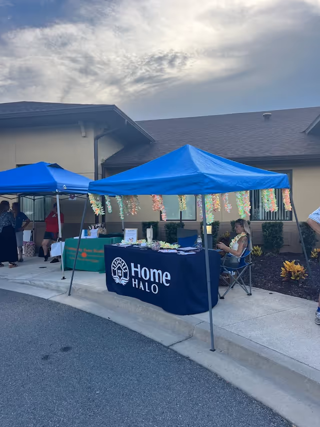 Outdoor event with blue canopies, a Home Halo table, and attendees outside a facility.