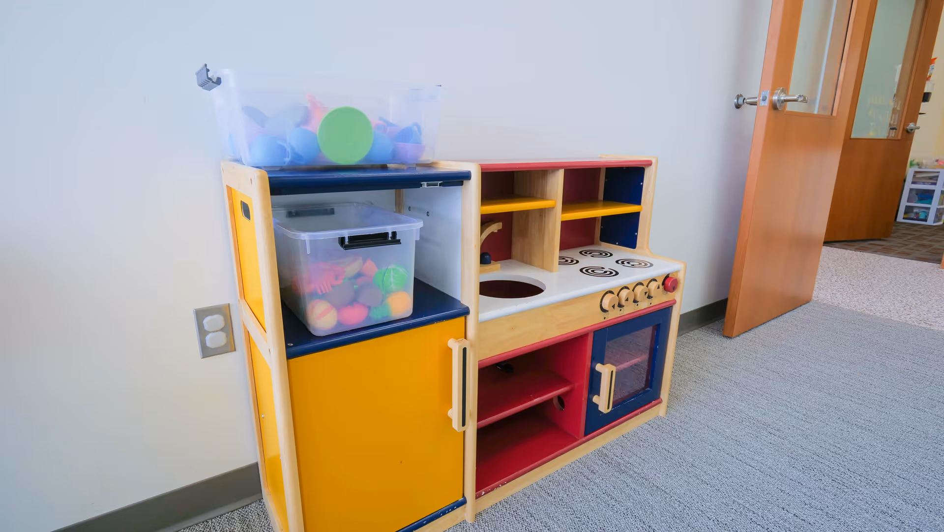 Colorful wooden toy kitchen playset with shelves sits in a carpeted bright room.