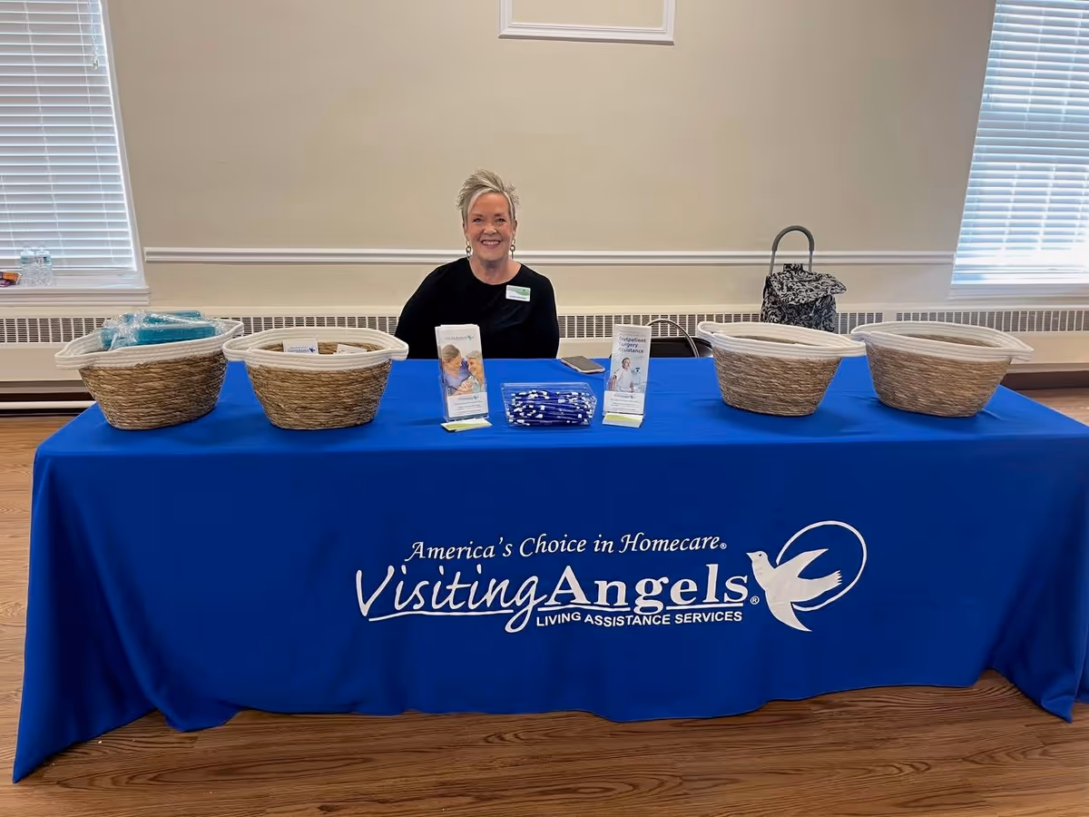 Smiling staff member sits behind a blue table displaying Visiting Angels materials indoors.