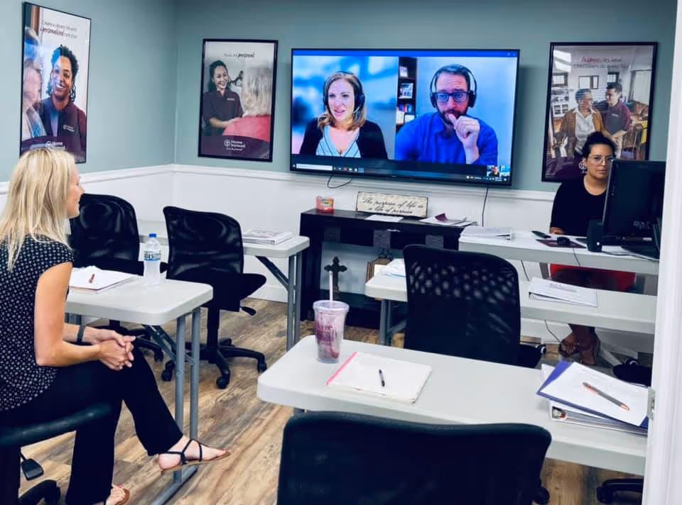 Home Instead training room with desks, chairs, and a video conference on a wall screen.