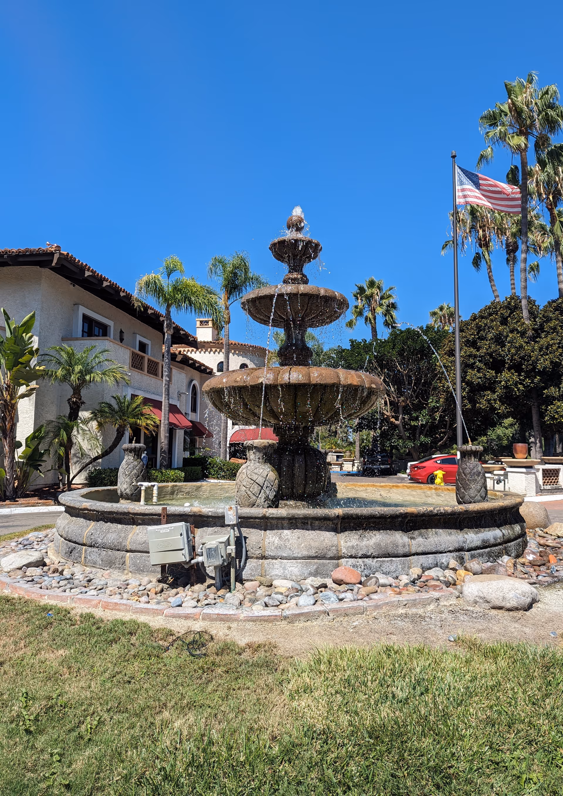 Outdoor courtyard fountain with palm trees and flag near a beige facility building.