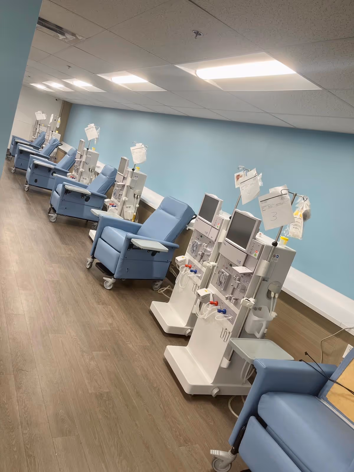 Row of blue dialysis chairs with machines in a clinical treatment room.