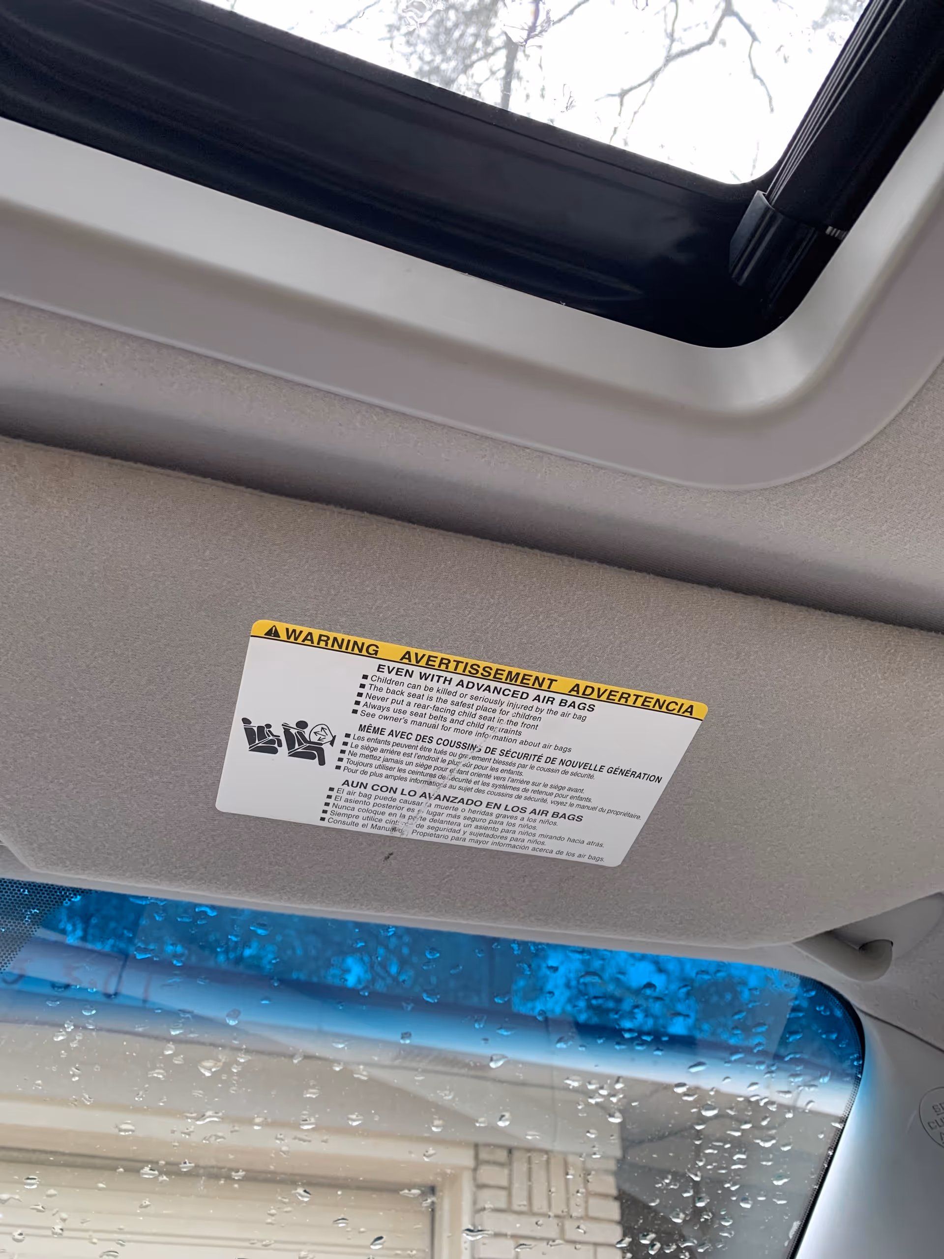 Car ceiling with warning label about airbags and a sunroof, raindrops on the window.
