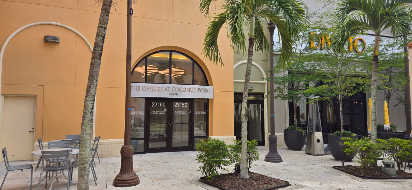 Outdoor plaza with palm trees, tables, chairs, and storefronts along a walkway.