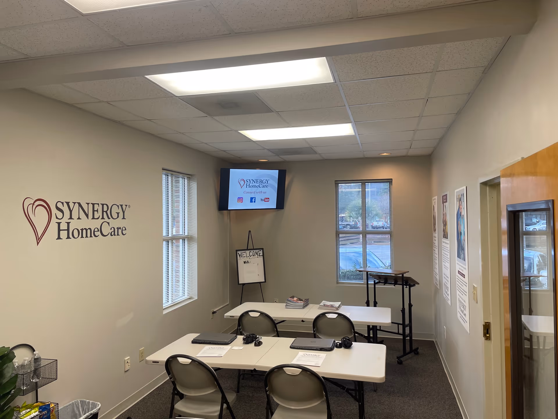 Interior reception area with SYNERGY HomeCare logo, tables, chairs, and a wall-mounted screen.