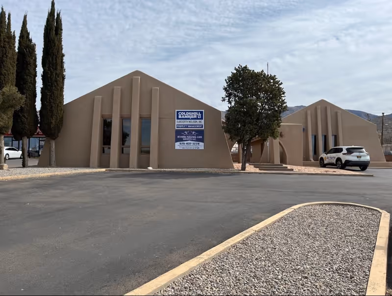 Beige angular building with a sign, trees, and a parked white SUV in front.