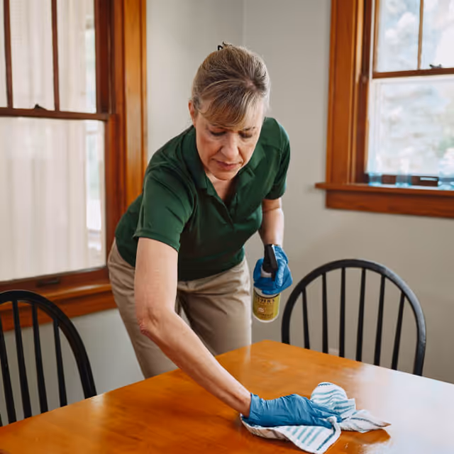 A caregiver in a green shirt cleans a wooden dining table indoors.