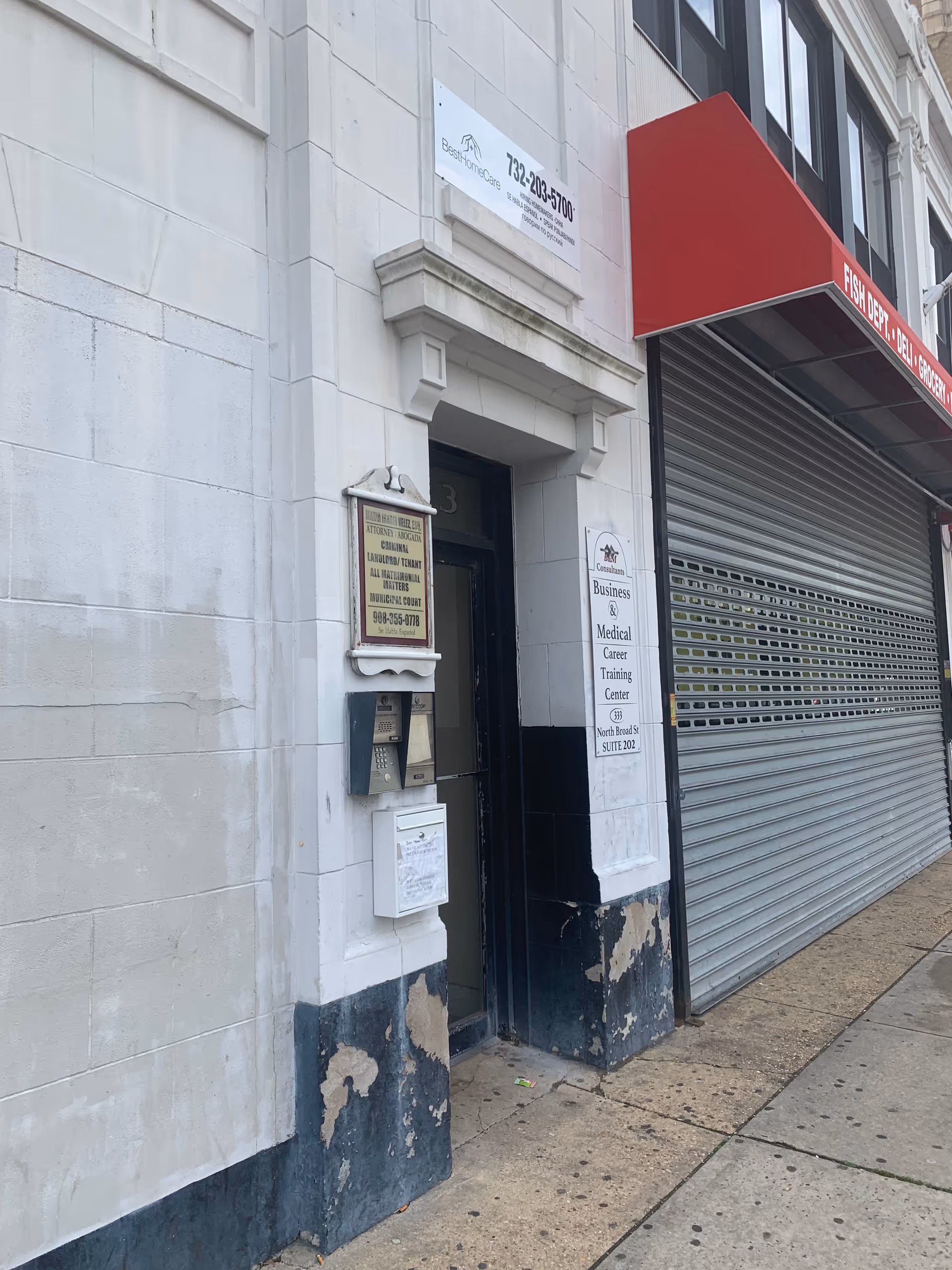 Urban building exterior with an entrance, signage boards, and a red awning beside a metal shutter.