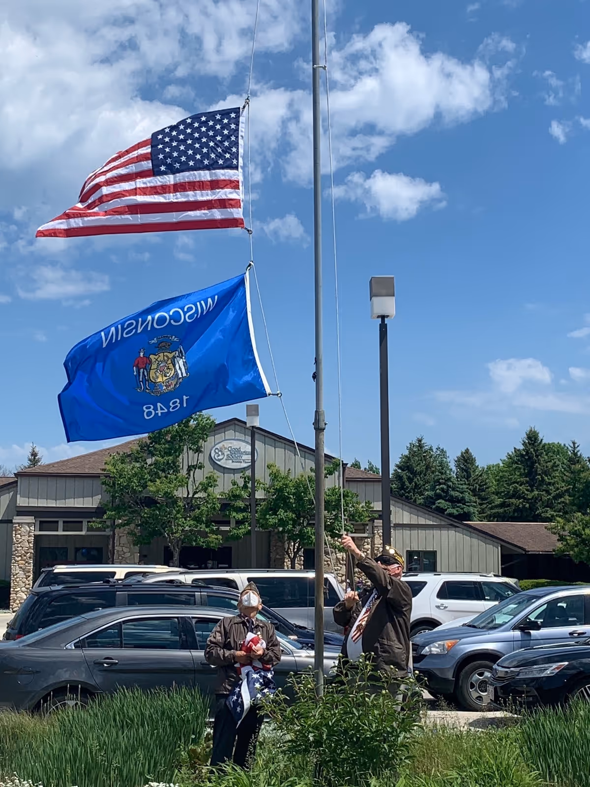 Two masked individuals raise US and blue state flags outside Unity Hospice building.