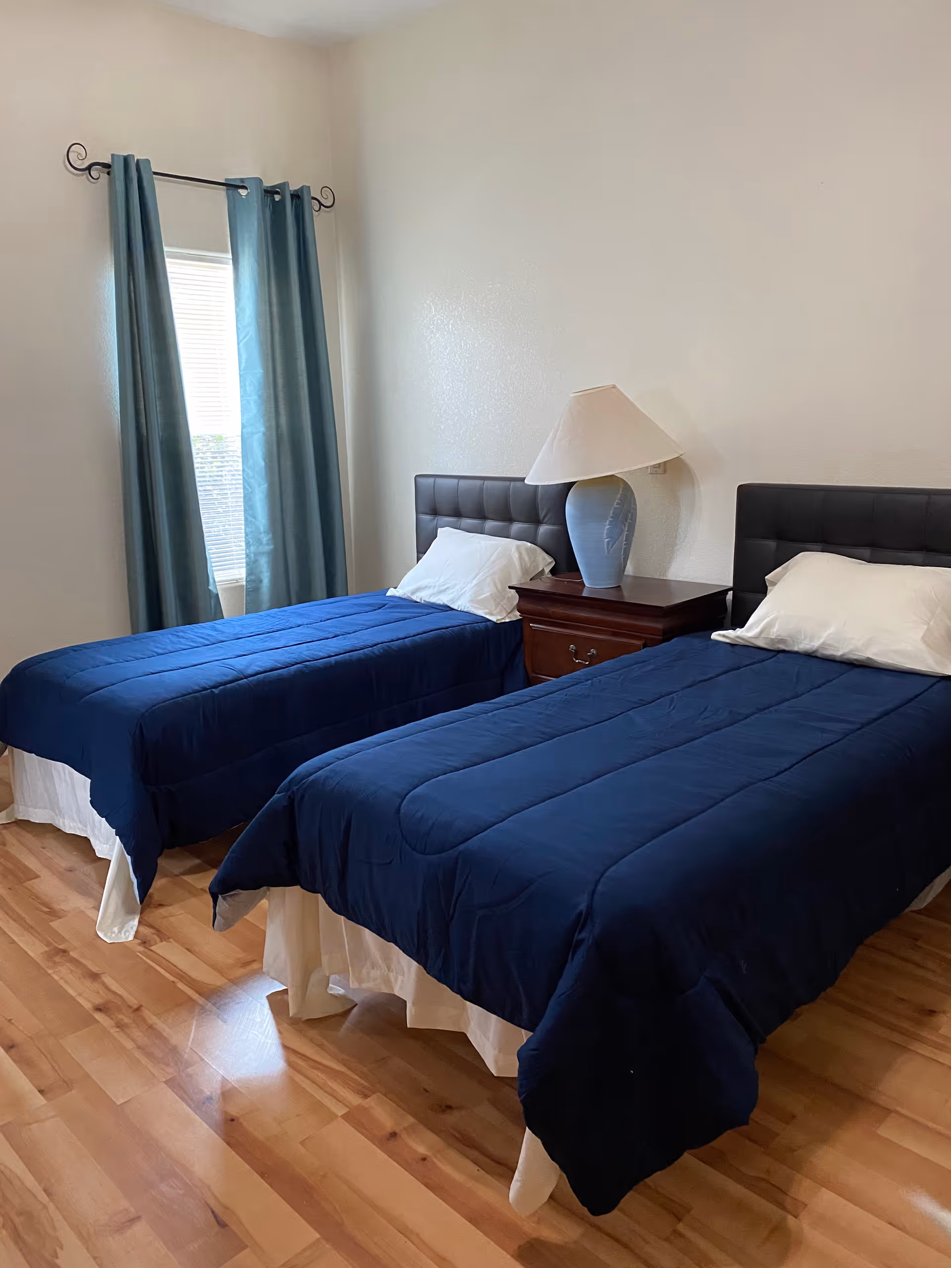 Two twin beds with blue quilts and a nightstand in a bright, simple bedroom.