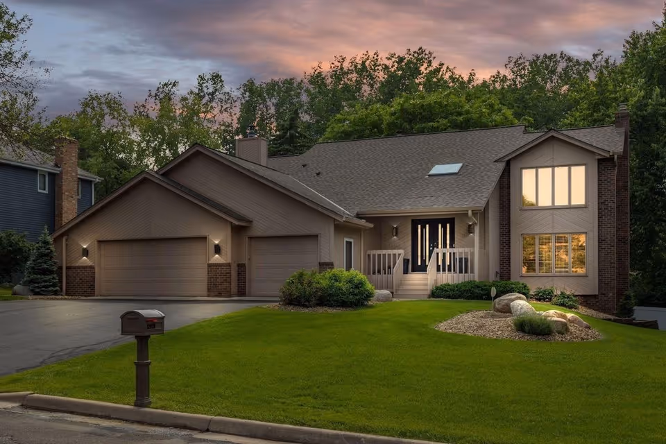 A modern suburban house with a three car garage, manicured lawn, and rock garden in front.
