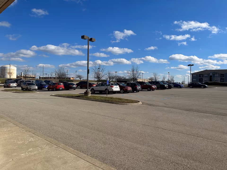 A spacious parking lot with numerous cars in front of a low-rise building under blue sky.