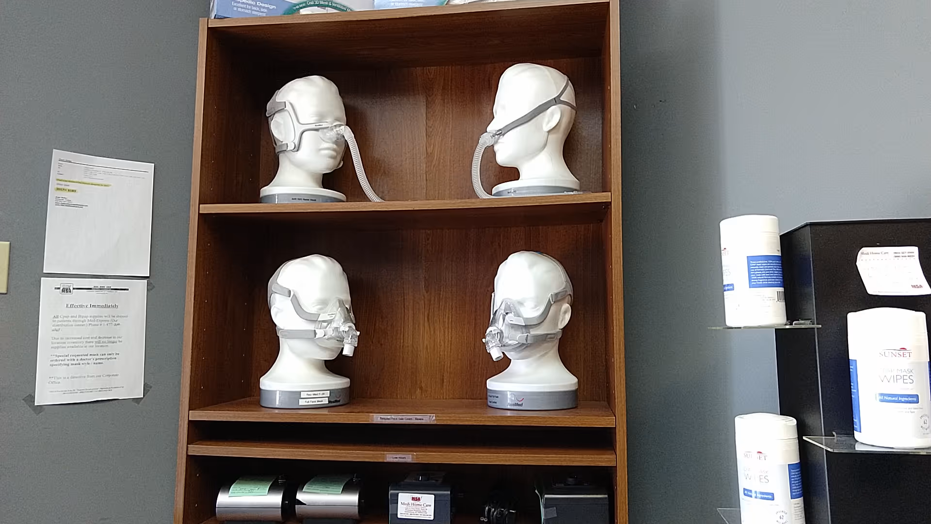 Wooden shelving holds CPAP mask mannequin heads and medical supplies in an interior facility room.