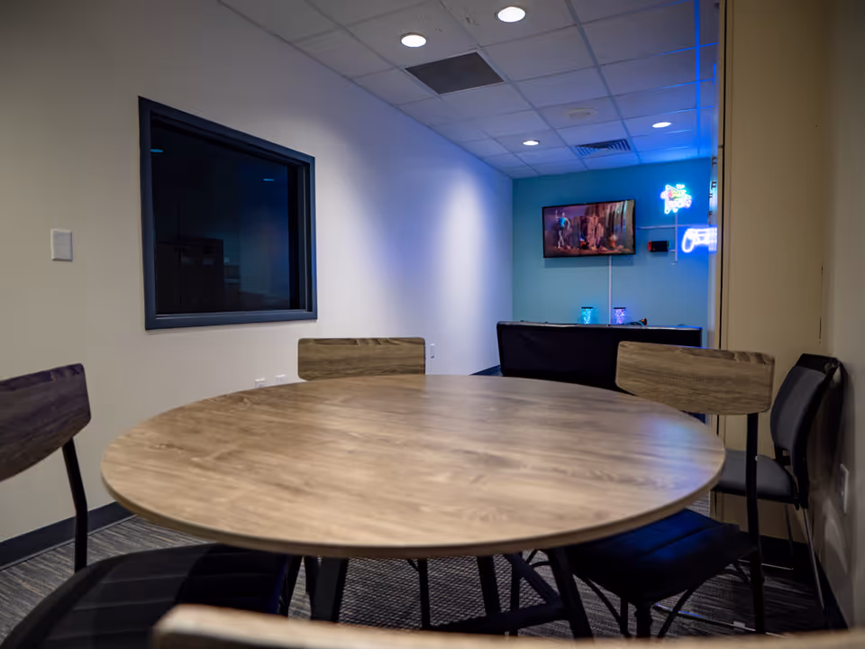 Round wooden table with chairs in a lounge-like interior, TV on blue wall.