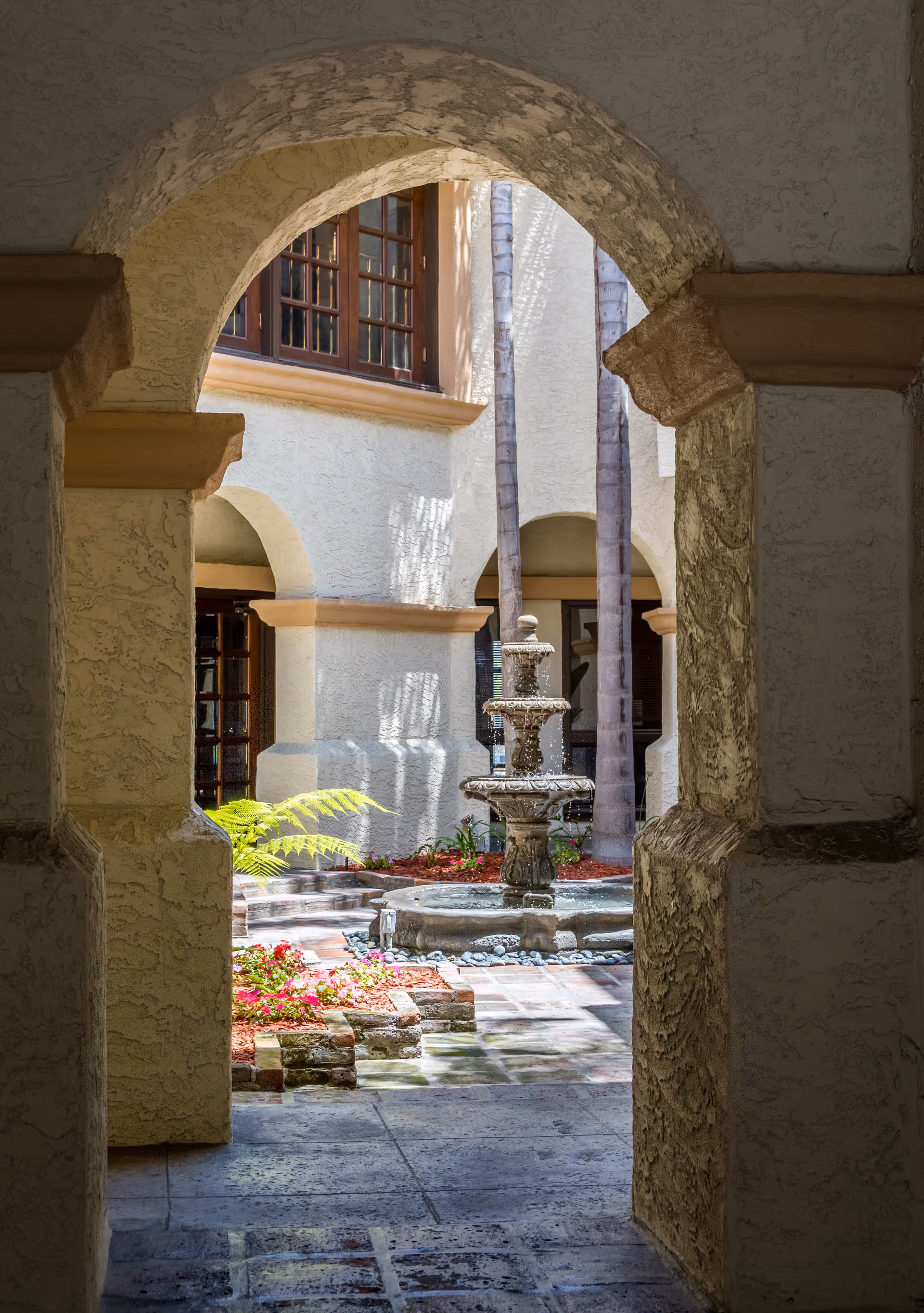 Architectural arch frames a sunlit courtyard with a central fountain and plants.