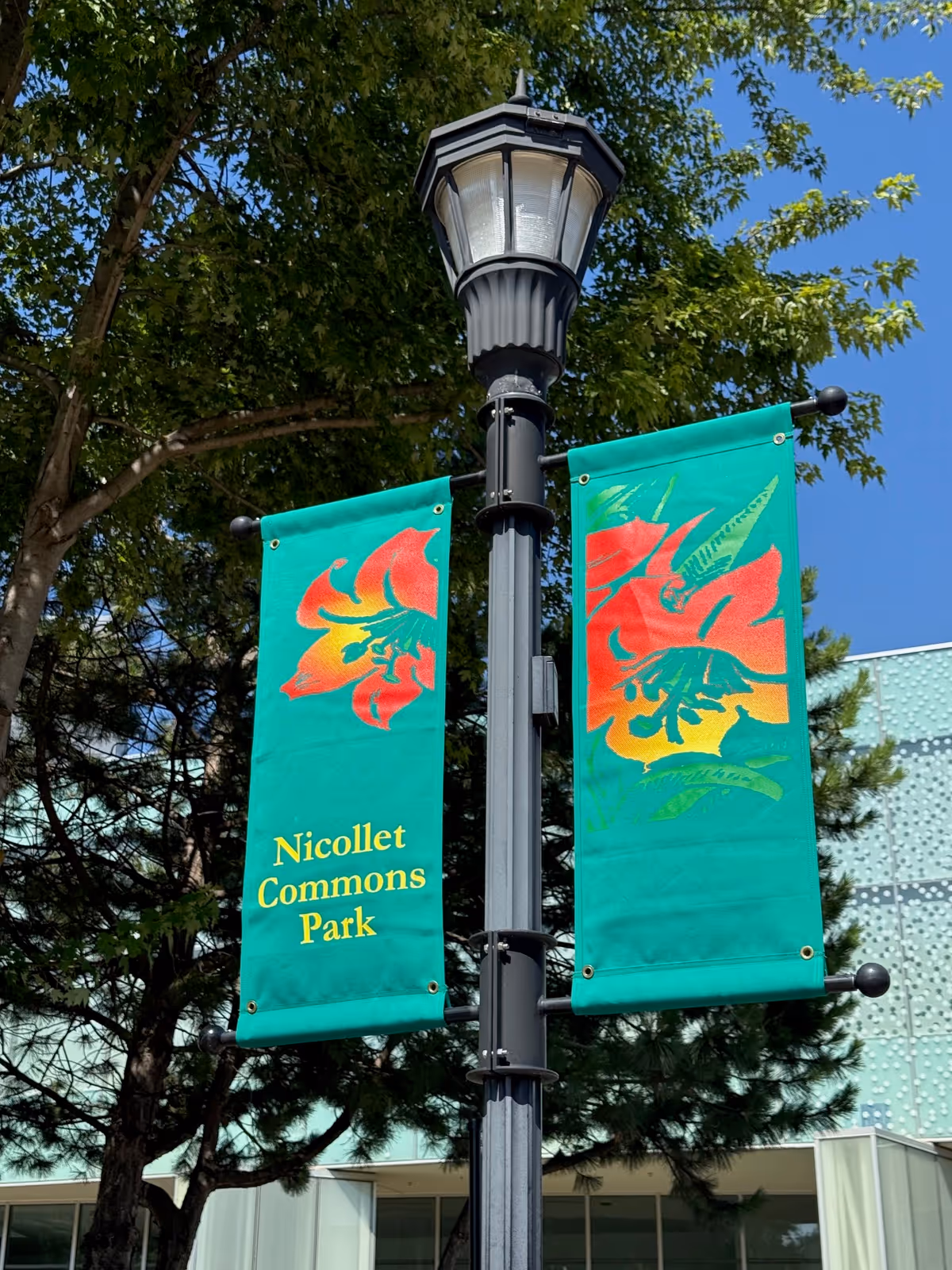 A black street lamp with green banners reading Nicolet Commons Park against a blue sky.