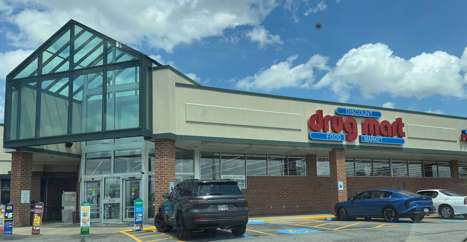 Exterior of Discount Drug Mart storefront with glass entry and parked cars.
